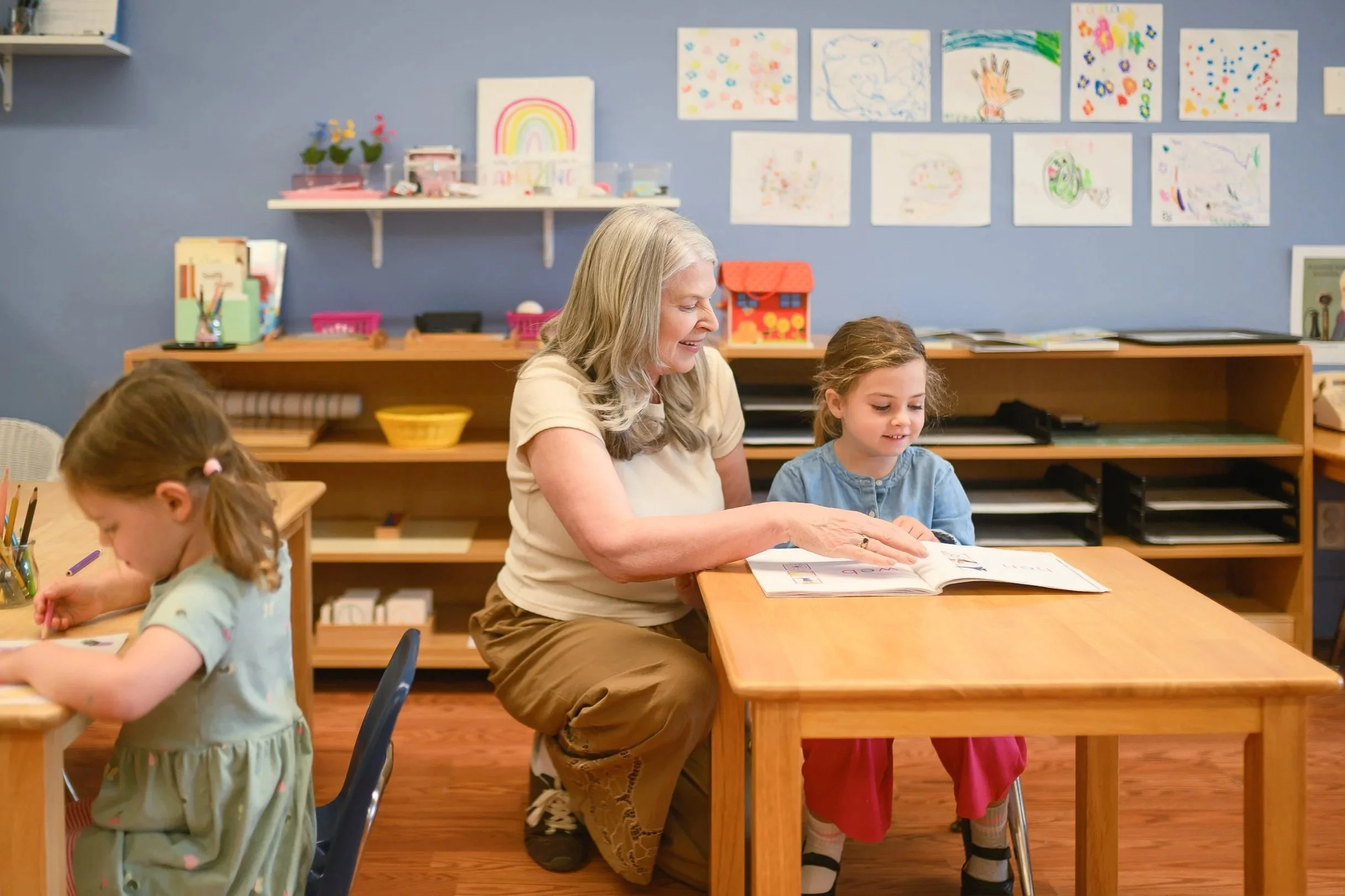 An elderly woman and two young girls sit at a wooden table in a classroom, engaging in reading or drawing activities. The background has a blue wall decorated with children's artwork, shelves with books, and colorful art supplies.
