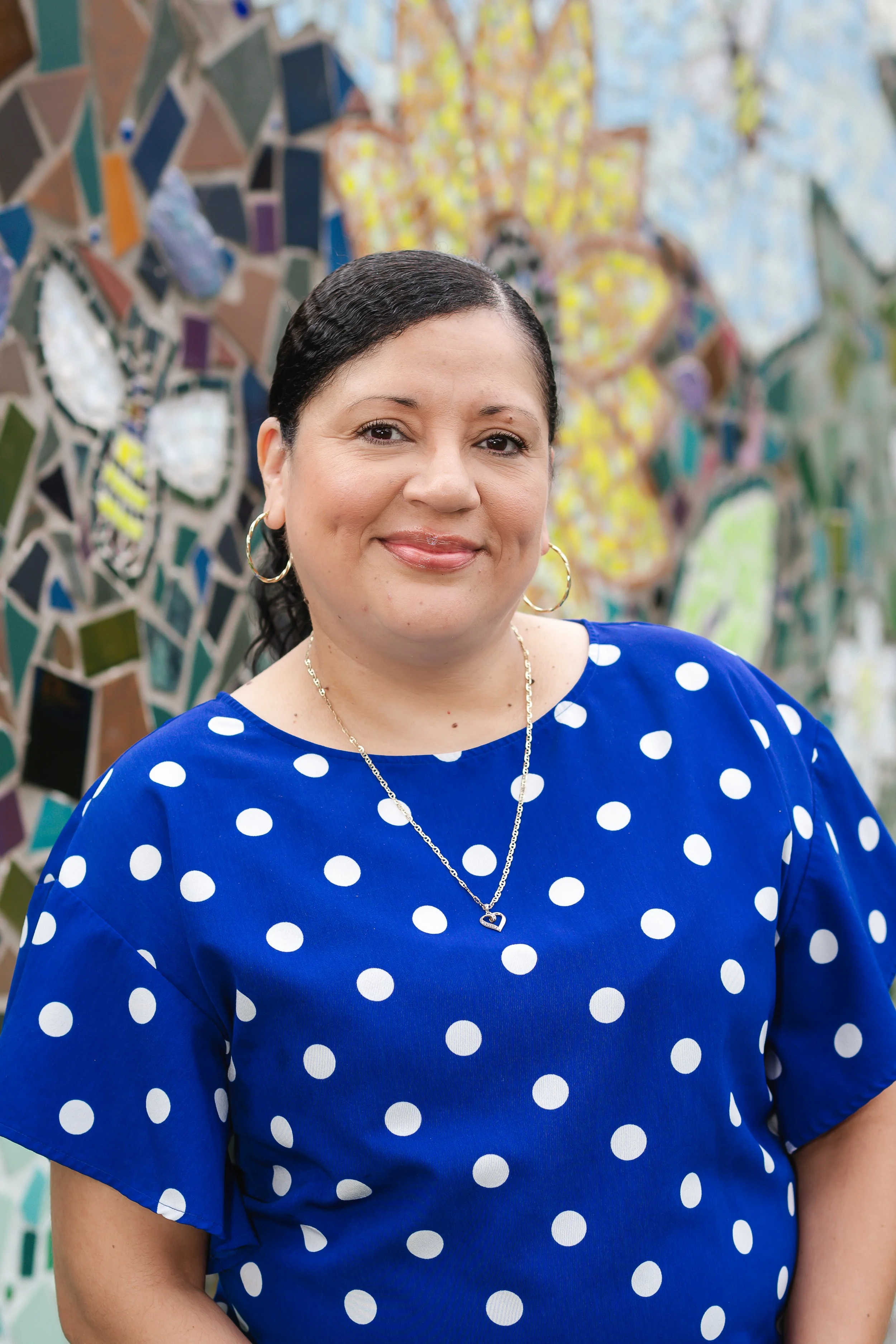 A woman with short dark hair, wearing a blue dress with white polka dots, gold hoop earrings, and a silver necklace with a heart-shaped pendant, stands in front of a colorful mosaic wall.