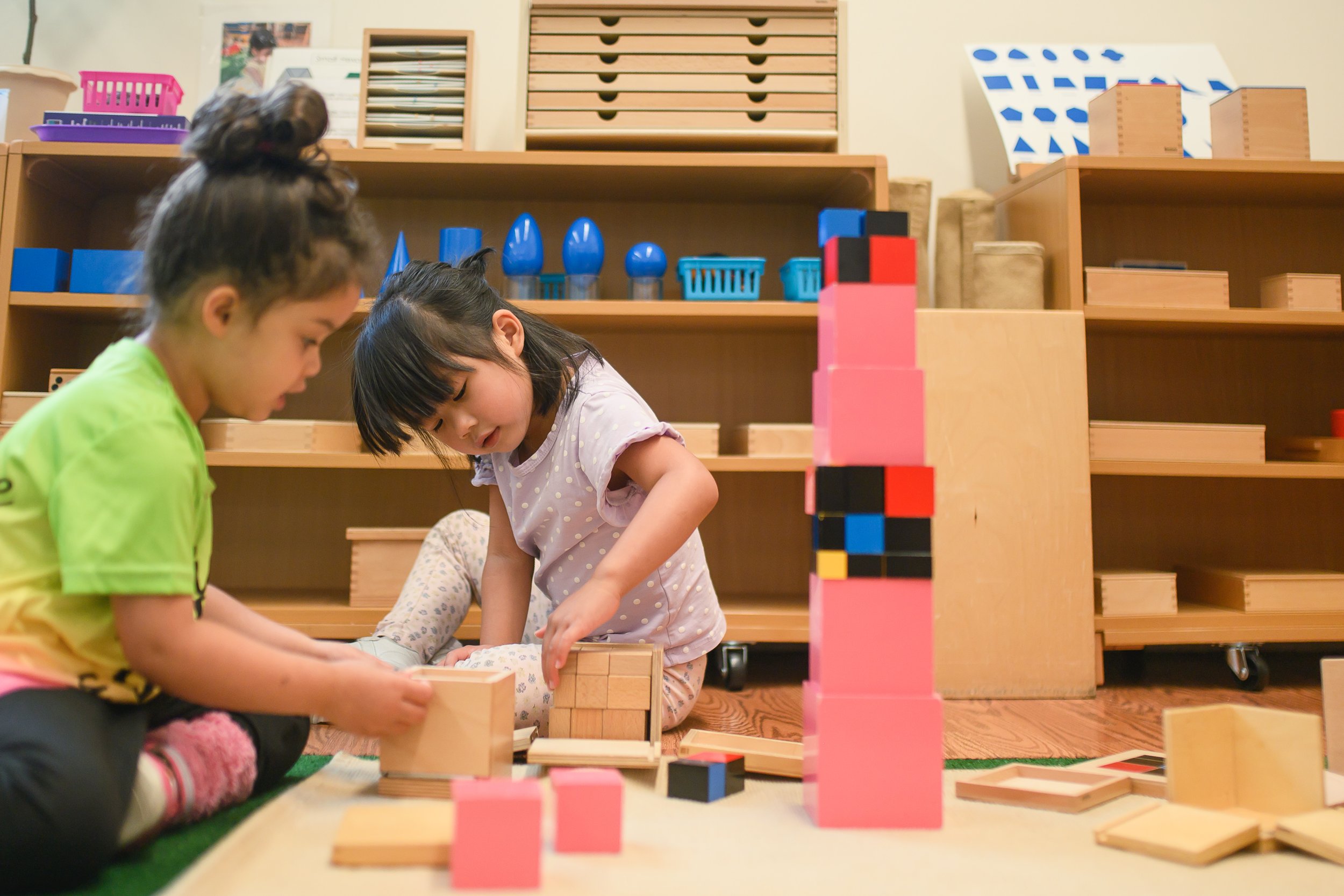 Two young girls playing with wooden blocks in a classroom or play area, with shelves containing various toys and educational materials in the background.