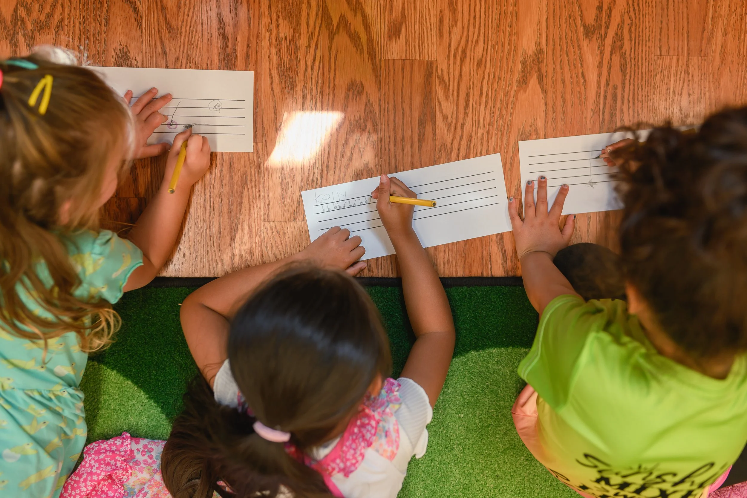 Children sitting on a green carpet at a wooden table, drawing on sheets of paper with yellow pencils.