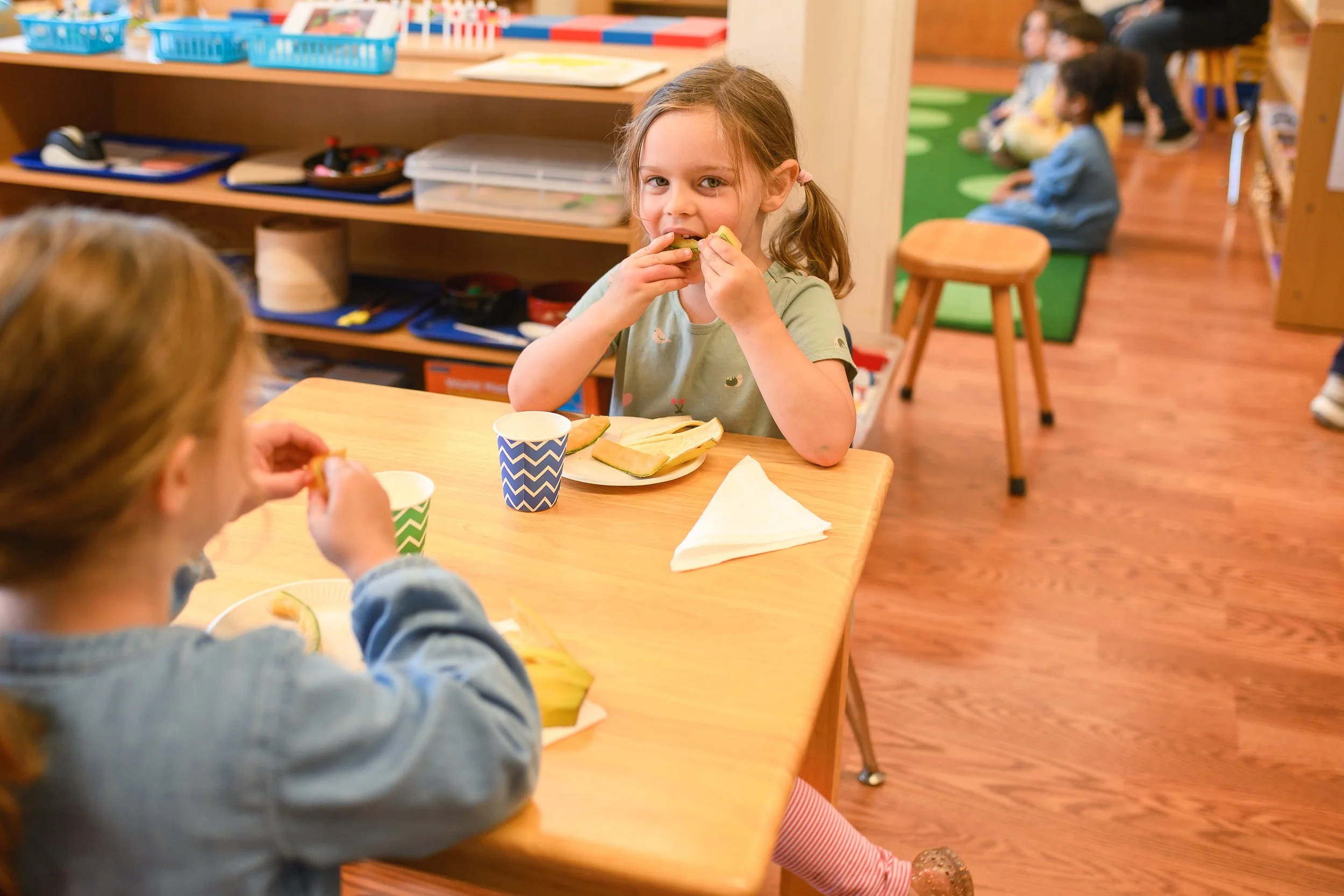 A young girl with brown hair in pigtails eating a sandwich at a table in a classroom.