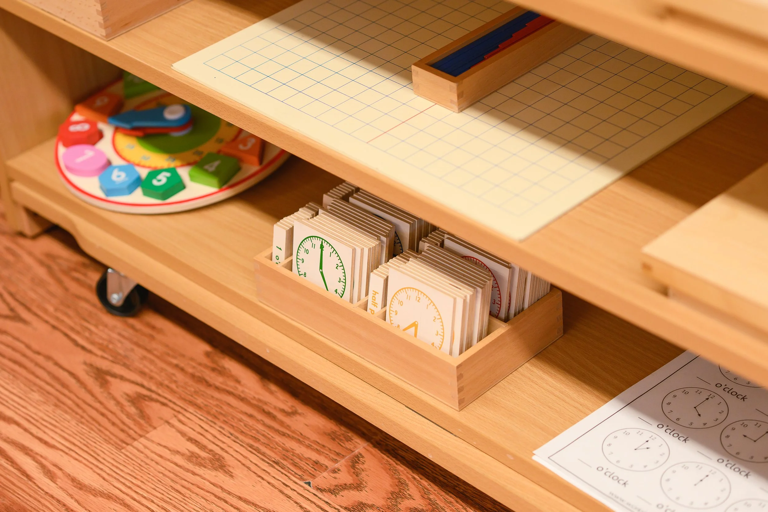 A wooden shelf with various educational tools including colorful clock puzzle pieces, time-telling cards, and a large worksheet with clock illustrations.