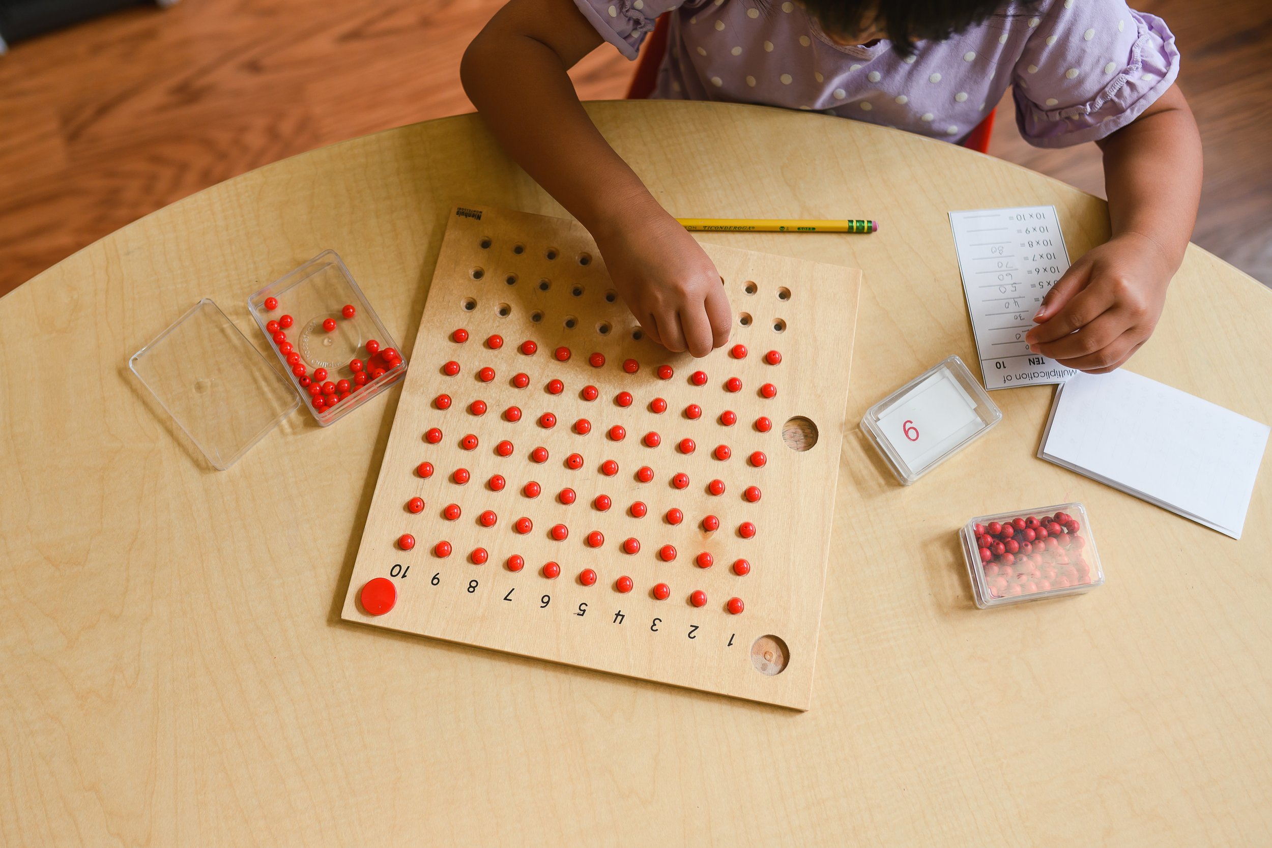 A child wearing a polka dot shirt is working on a math activity at a light-colored wooden table. The activity involves a wooden pegboard with numbered holes, red plastic pegs, and a sheet of paper with multiple multiplication equations. The child appears to be placing the pegs into the pegboard to match the multiplication facts.