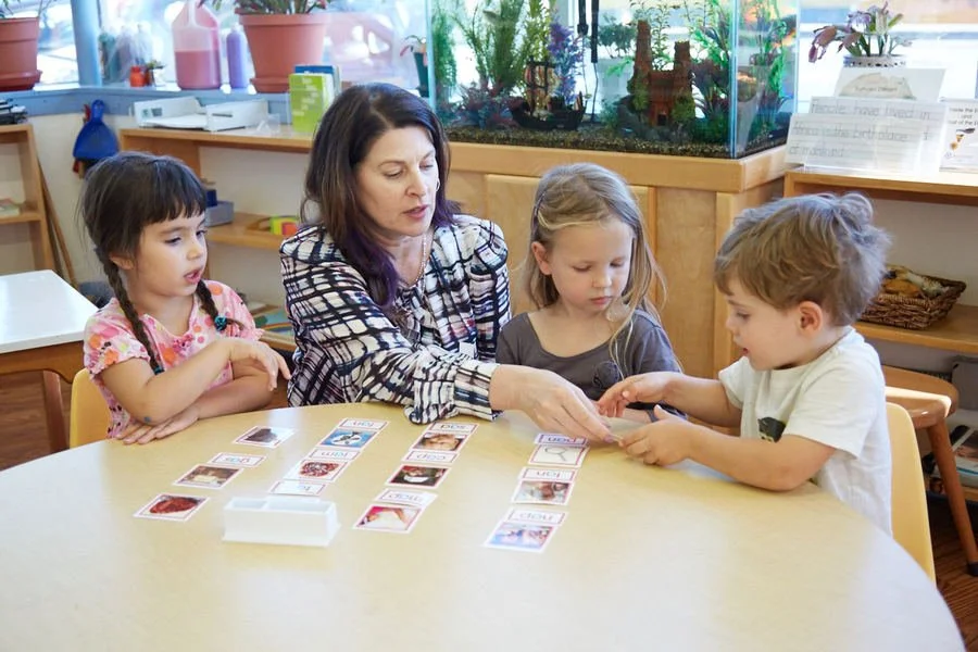 A teacher with three young children playing a card game at a table in a classroom.