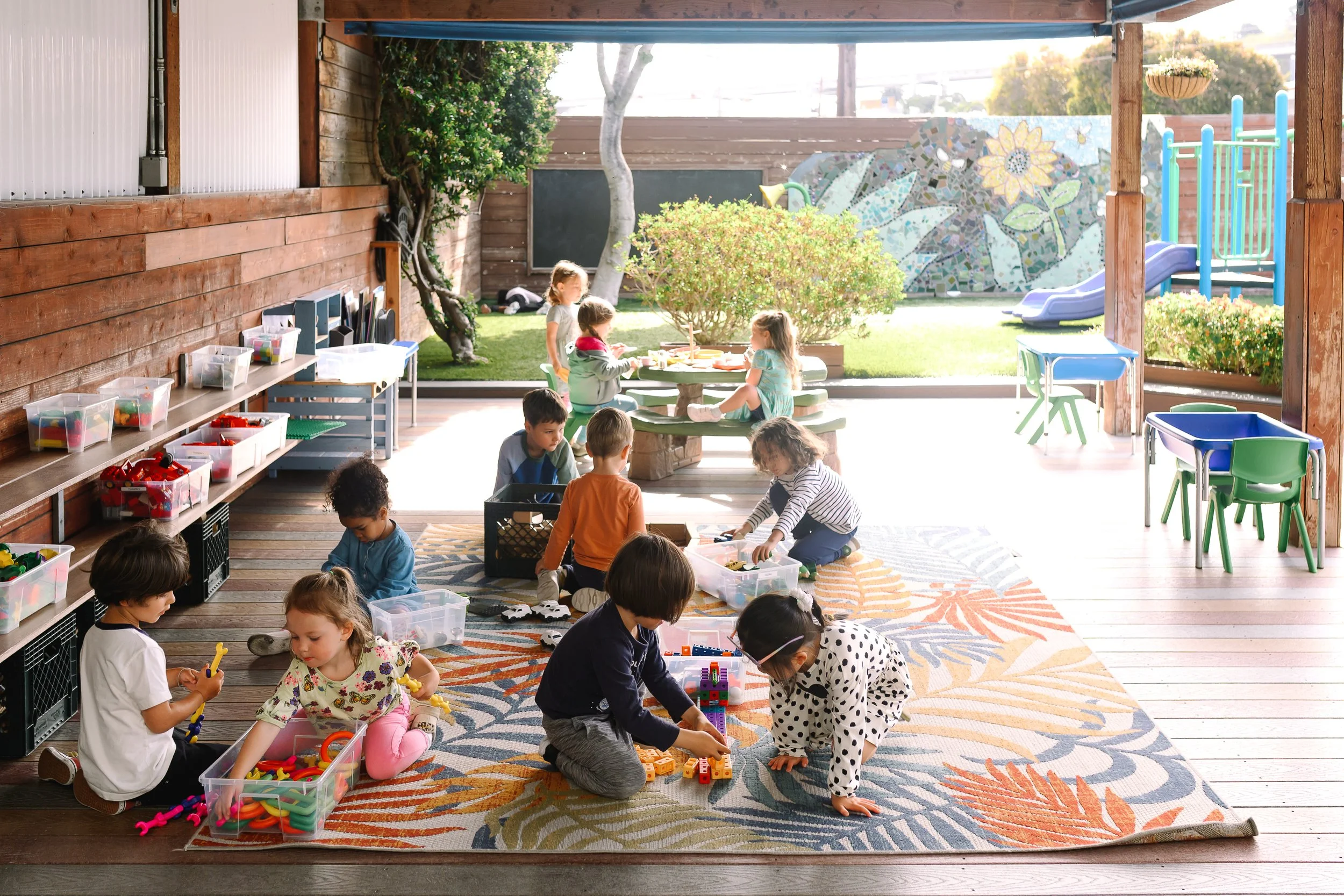 Children playing and building with colorful toys and blocks on a patterned rug in an outdoor covered area of a preschool or daycare.