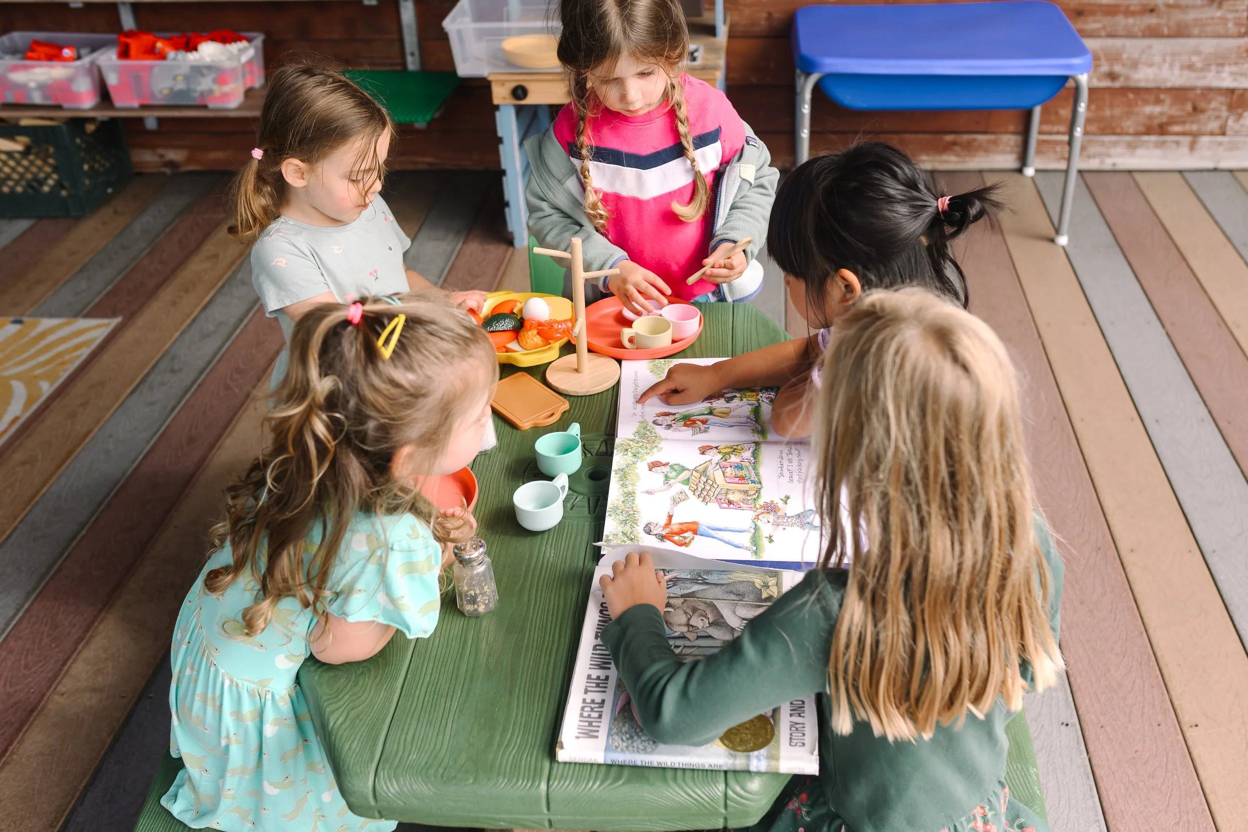 Group of young girls playing at a table with toy food and a storybook in a playroom.