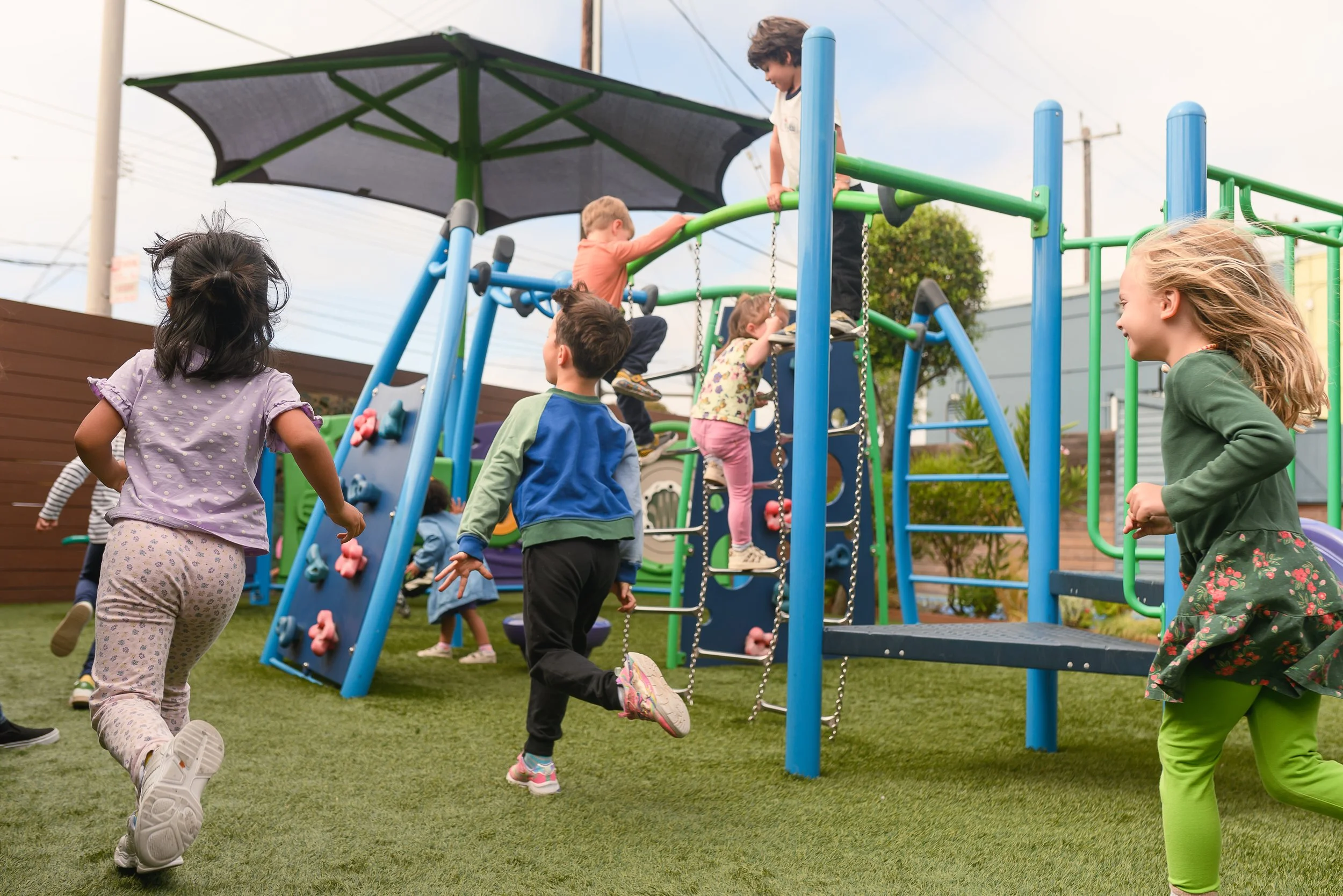 Children playing and climbing on a colorful playground set outdoors with a large umbrella and a wooden fence in the background.