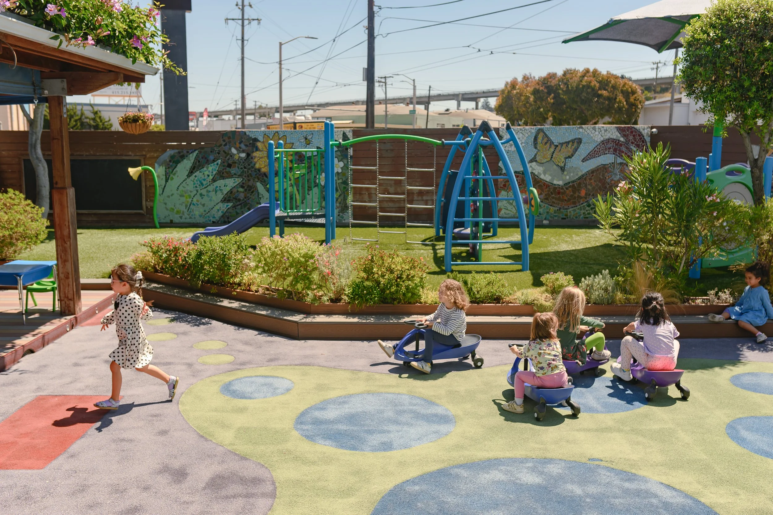 A colorful playground with children playing and sitting on circular step-like platforms, a slide, and various play equipment near a garden and a mural with butterflies and flowers, under a blue sky.