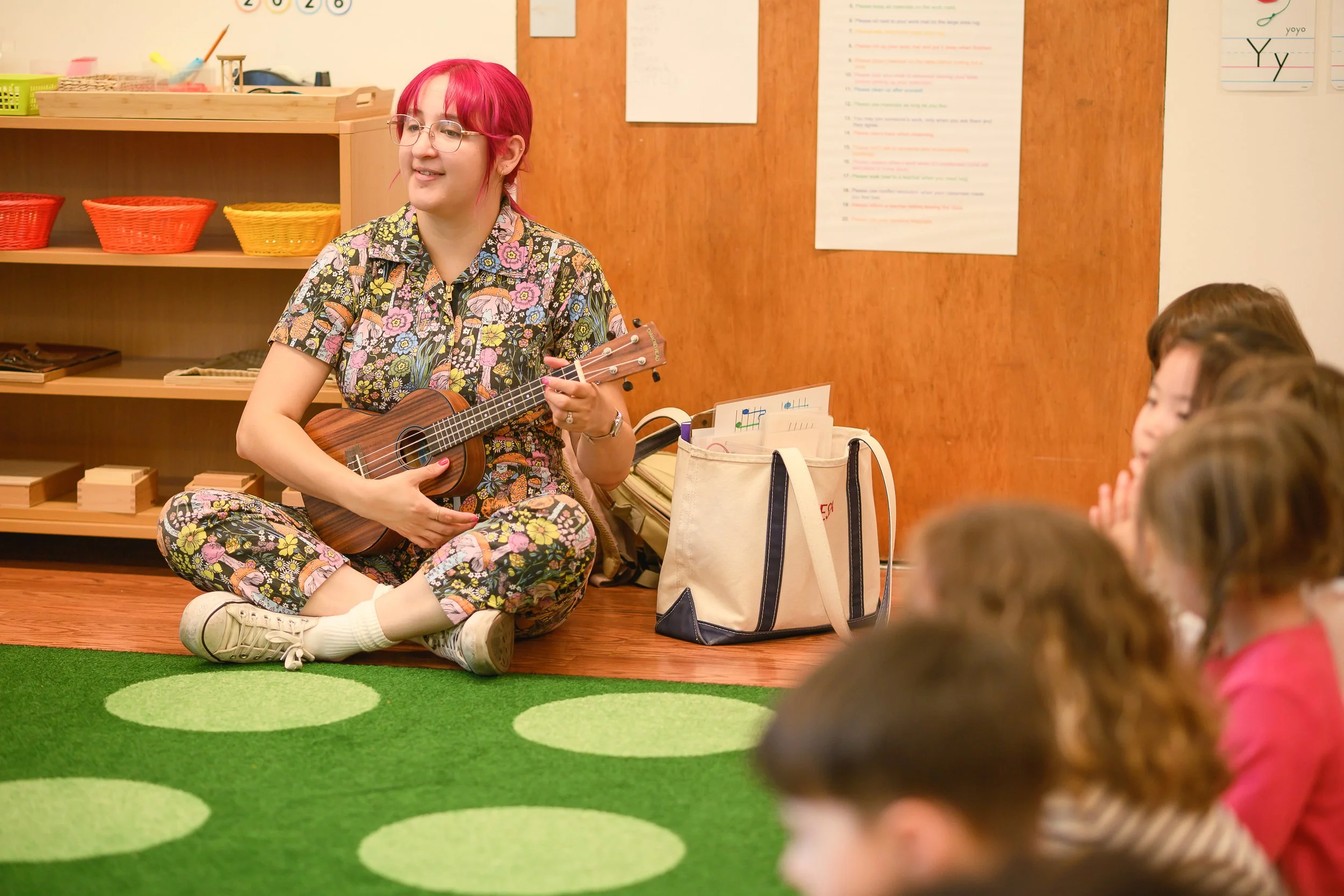 A woman with pink hair, glasses, and a floral patterned outfit is sitting on a green carpet with circular patterns, holding a ukulele, around children in a classroom.