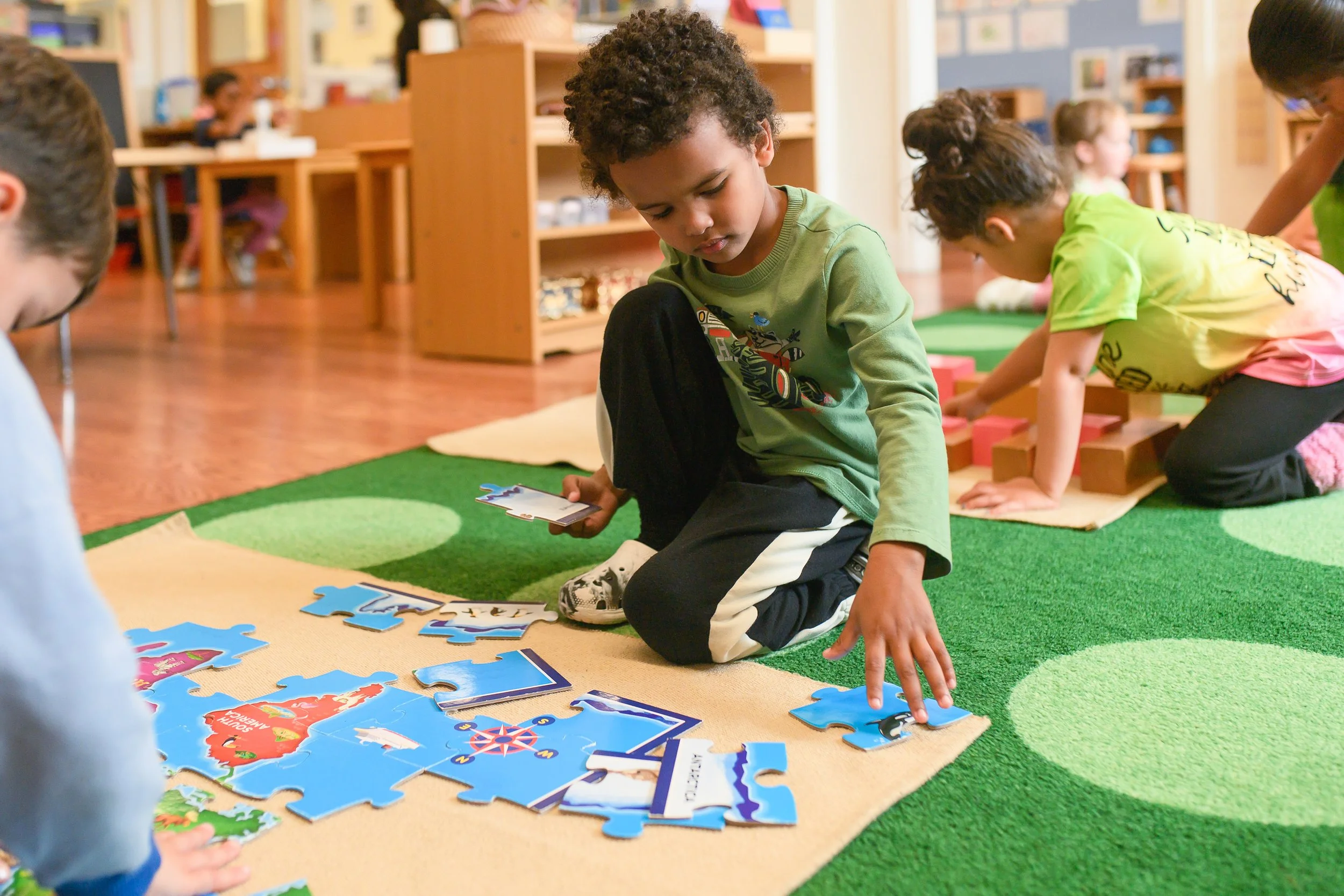 Children playing with a large puzzle on a colorful carpeted floor in a classroom.