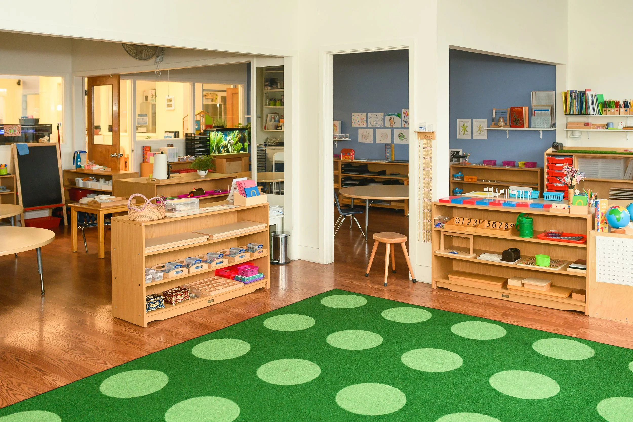 Interior of a preschool or daycare classroom with wooden furniture, colorful educational materials, and a bright green polka dot rug.