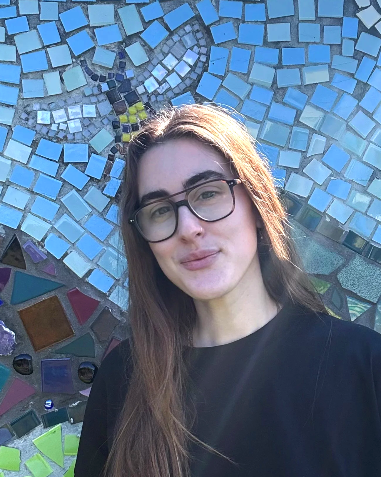 A young woman with long brown hair and glasses taking a selfie in front of a colorful mosaic wall with various geometric shapes and colors.