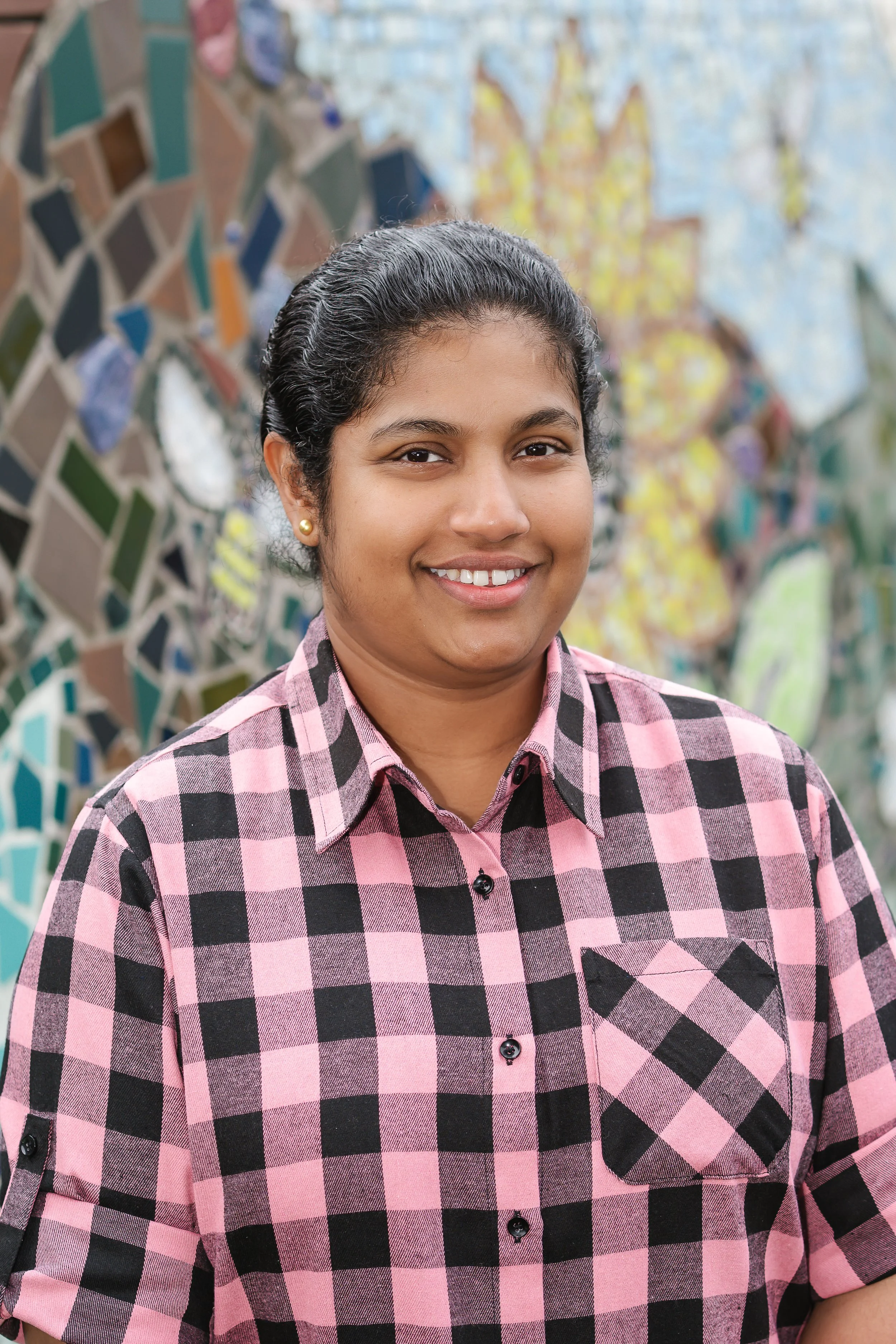A woman with dark hair in a ponytail, wearing a pink and black checkered shirt, smiling in front of a mosaic wall.
