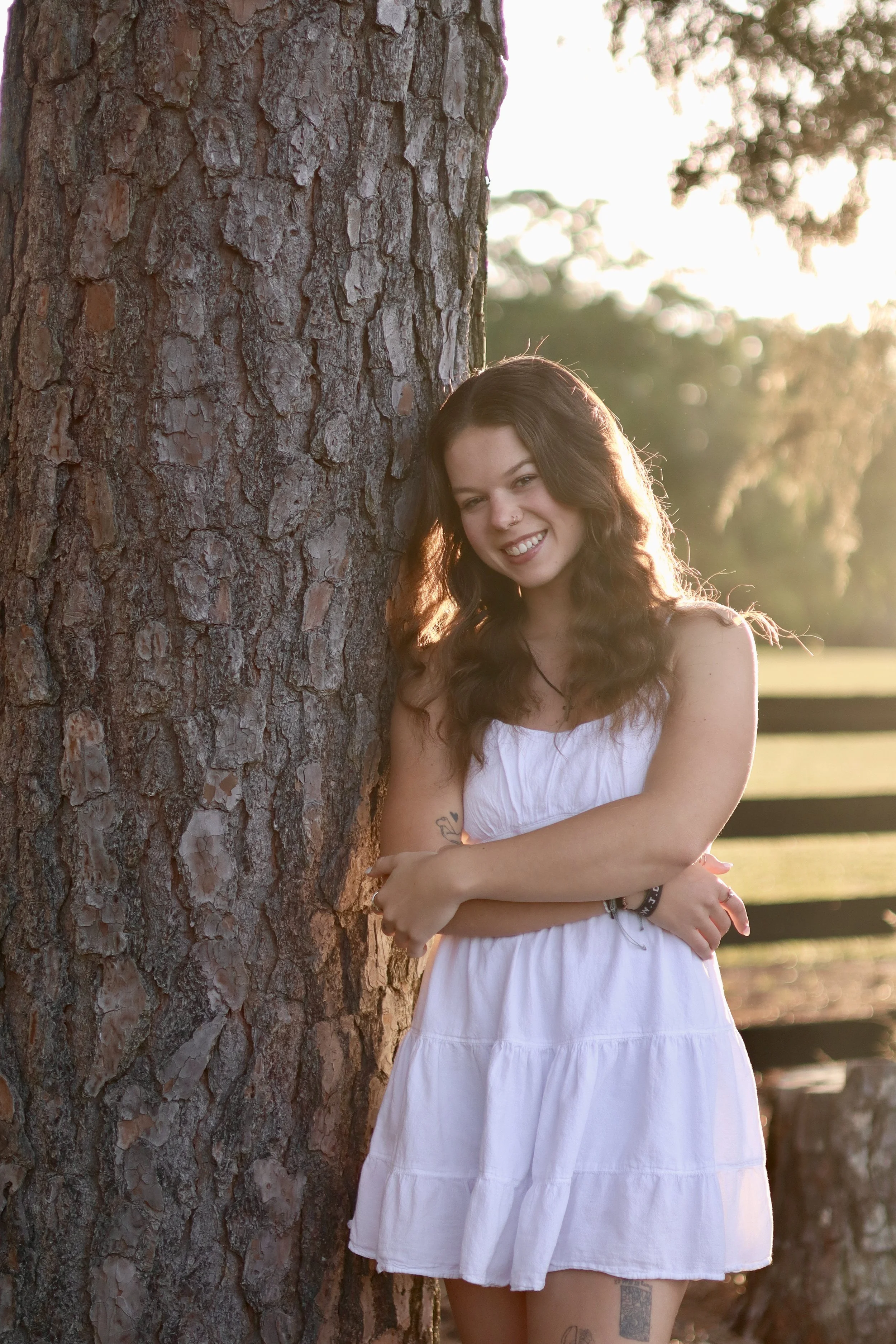 A young woman with dark, wavy hair, smiling, leaning against a tree with her arms crossed. She is wearing a white sundress and standing outdoors during sunset, with blurred trees and a wooden fence in the background.