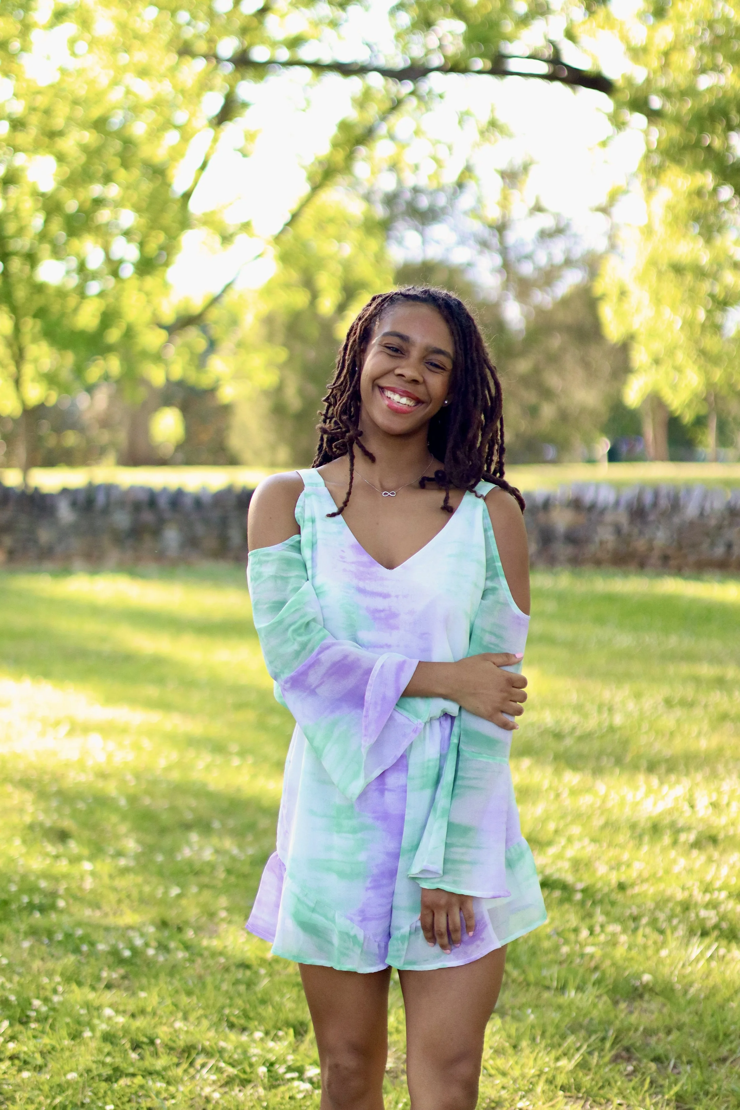 Young woman with dreadlocks smiling outdoors in a park with green trees and grass, wearing a pastel-colored dress.