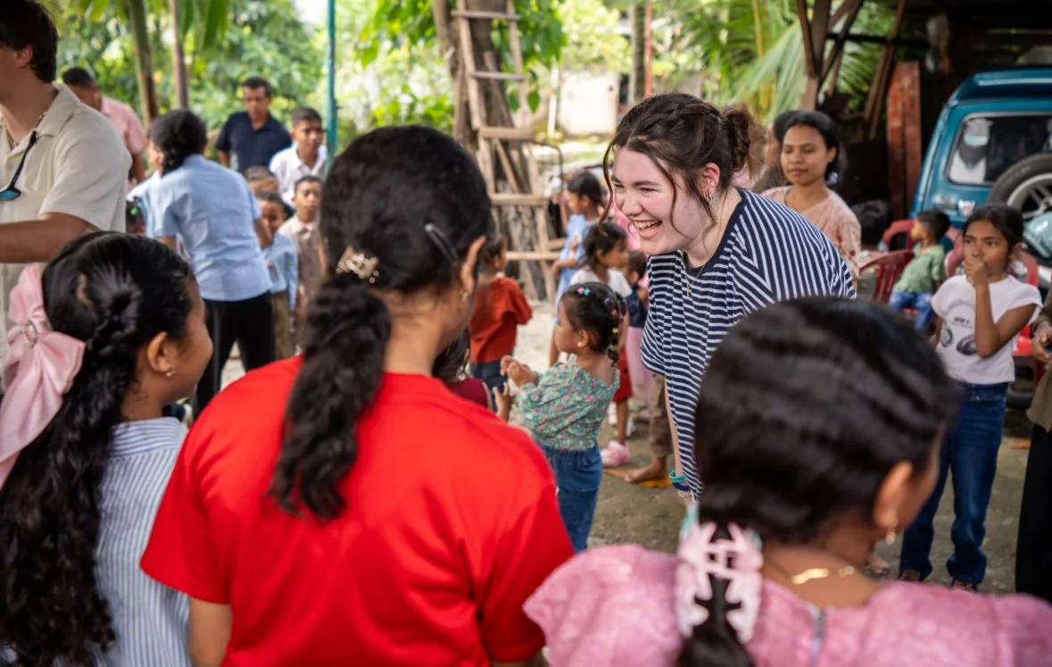 A woman with dark hair in a ponytail, wearing a striped shirt, smiles and leans forward while interacting with children at a community event outdoors, with other children and adults in the background.