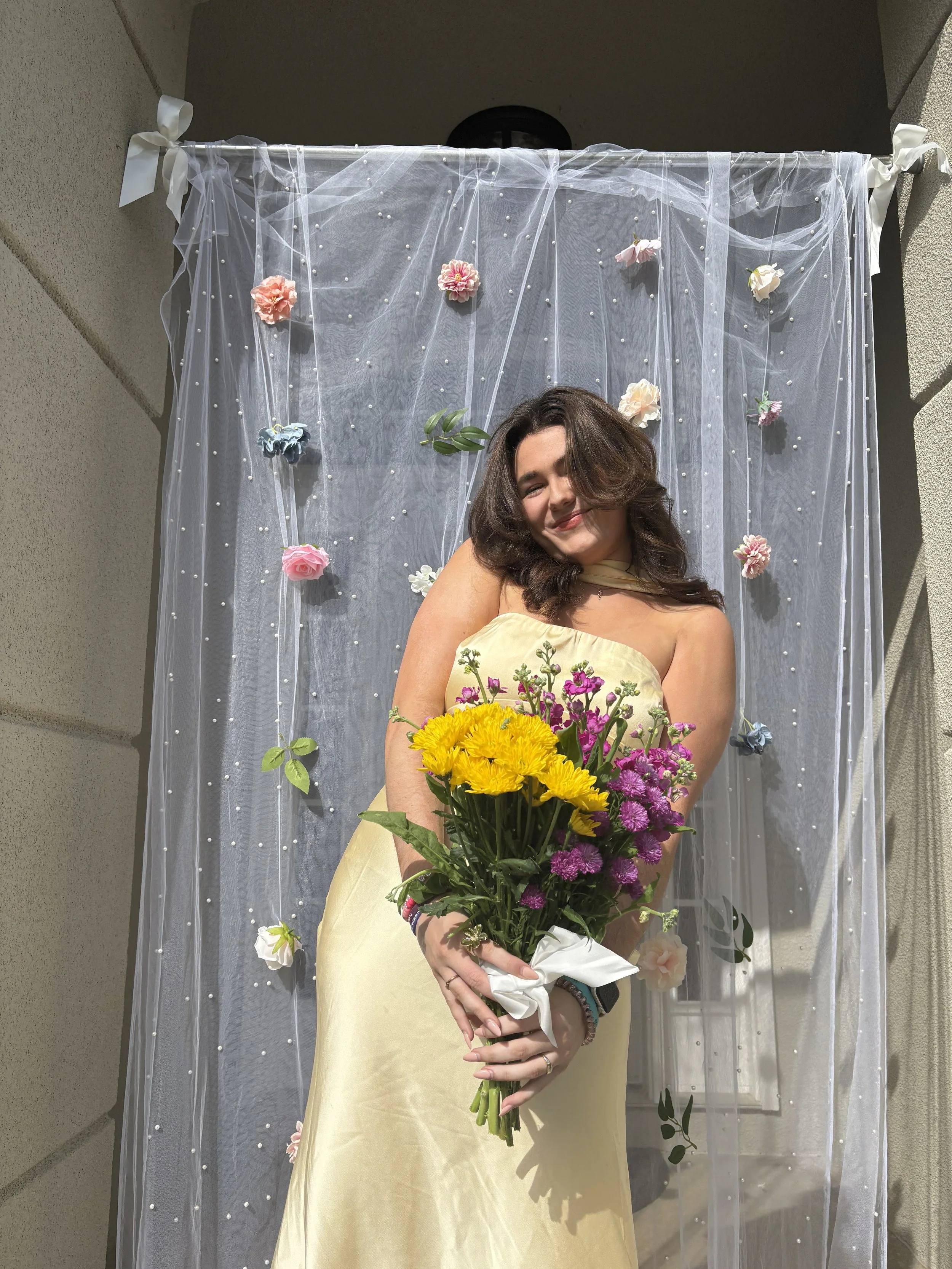 A woman in a light yellow strapless dress holding a large bouquet of yellow, purple, and pink flowers in front of a sheer white curtain decorated with flowers and pearls.