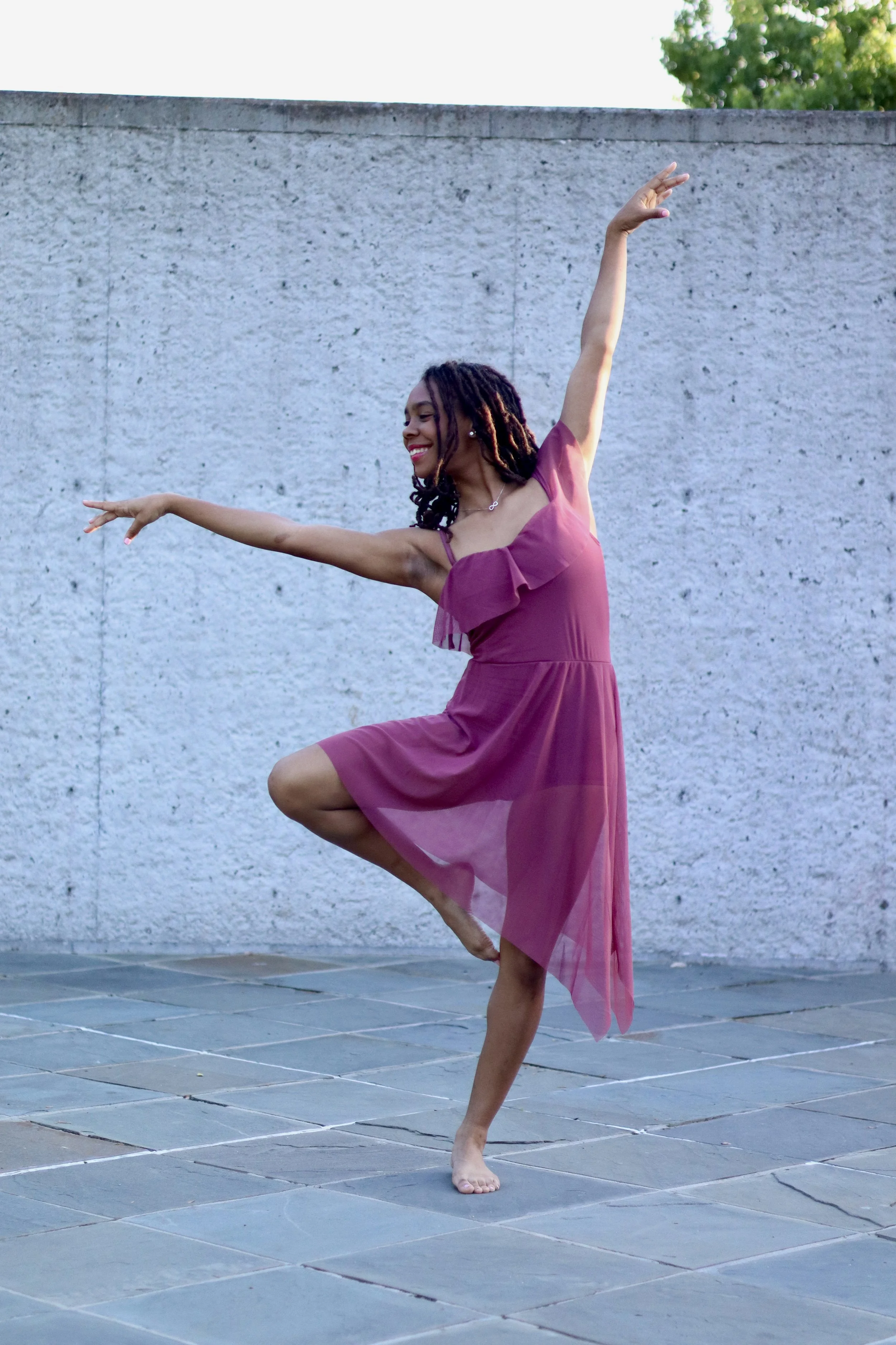 A woman with dark curly hair in a purple dress is balancing on one leg in a yoga pose outdoors on a paved surface, with a gray concrete wall and green trees in the background, smiling.