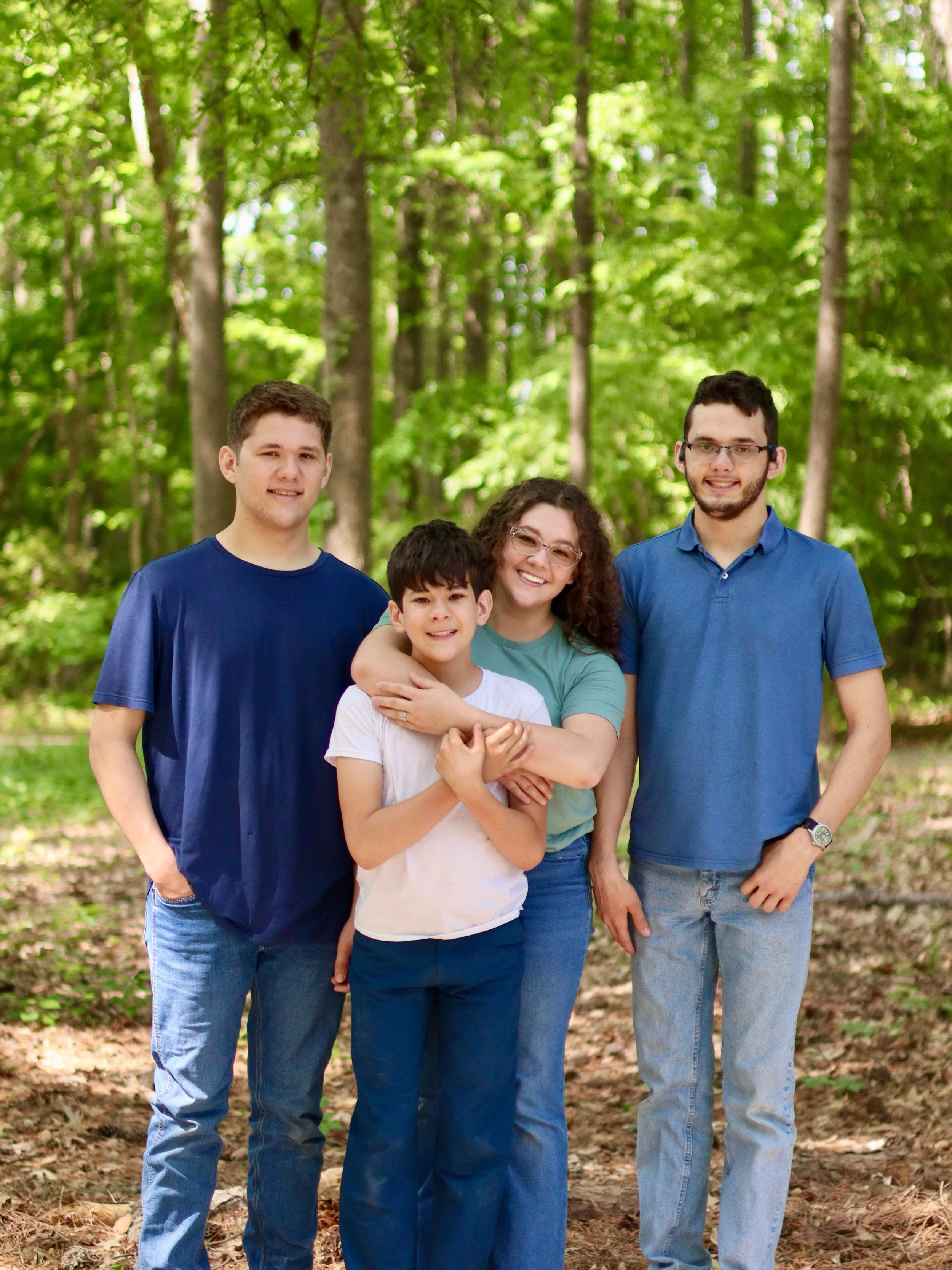 A group of five people standing outdoors in a forest, smiling and posing for a photo. There are three men and two women, with green trees and sunlight in the background.