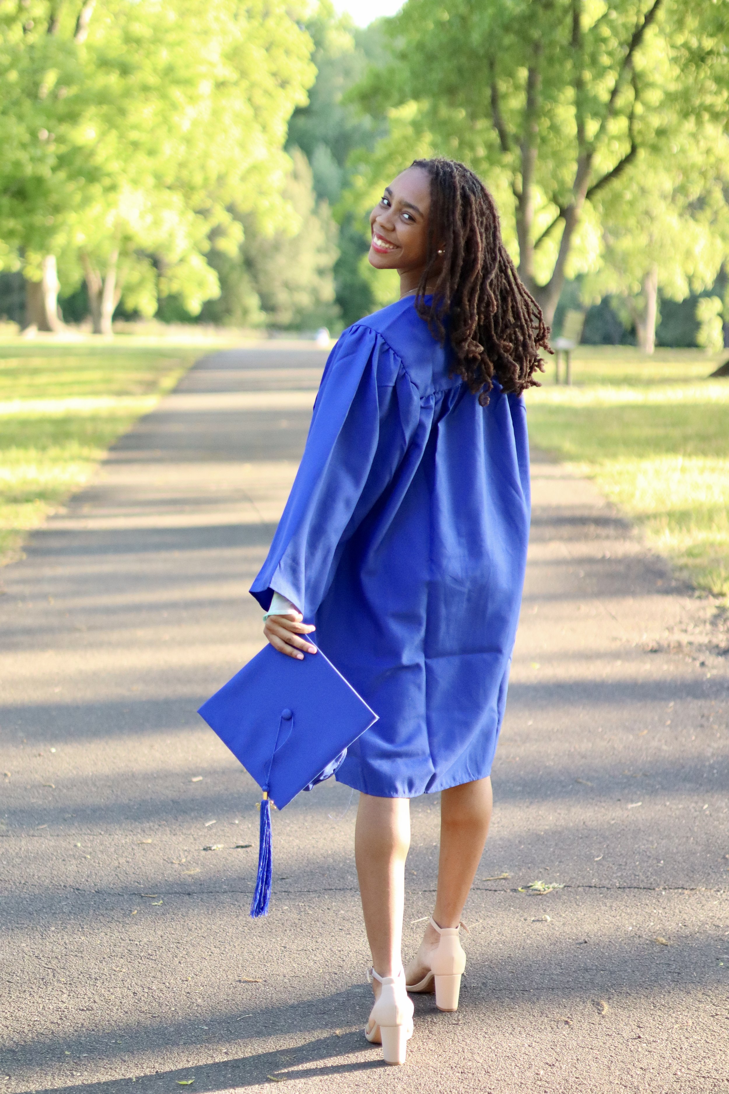 A young woman wearing a blue graduation gown and holding a matching blue cap with a tassel, walking on a paved path in a park with green trees and sunlight.