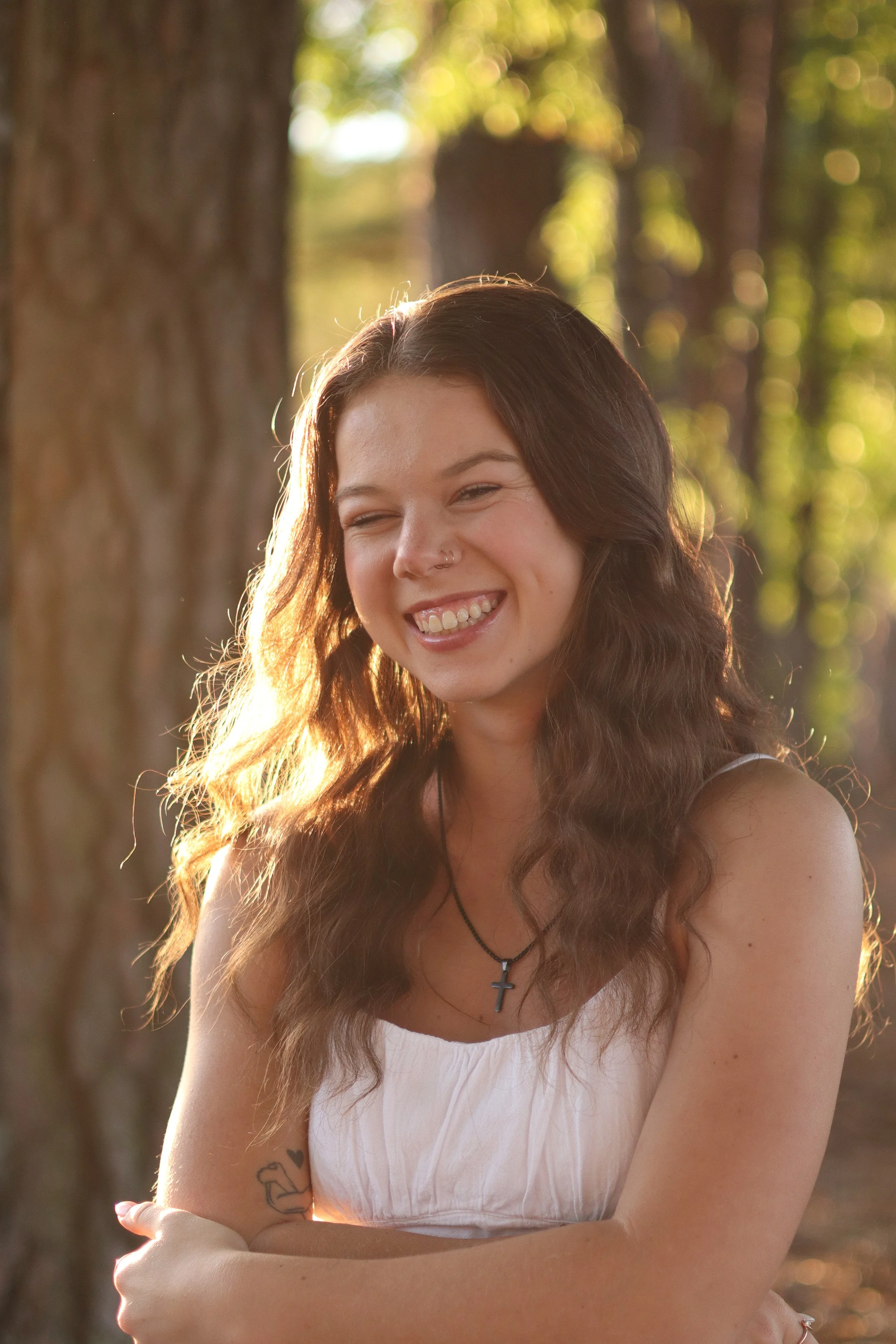 A young woman with long wavy hair and a nose piercing, smiling with her eyes closed, standing outdoors in front of trees illuminated by sunlight, wearing a white sleeveless top and a necklace with a cross.