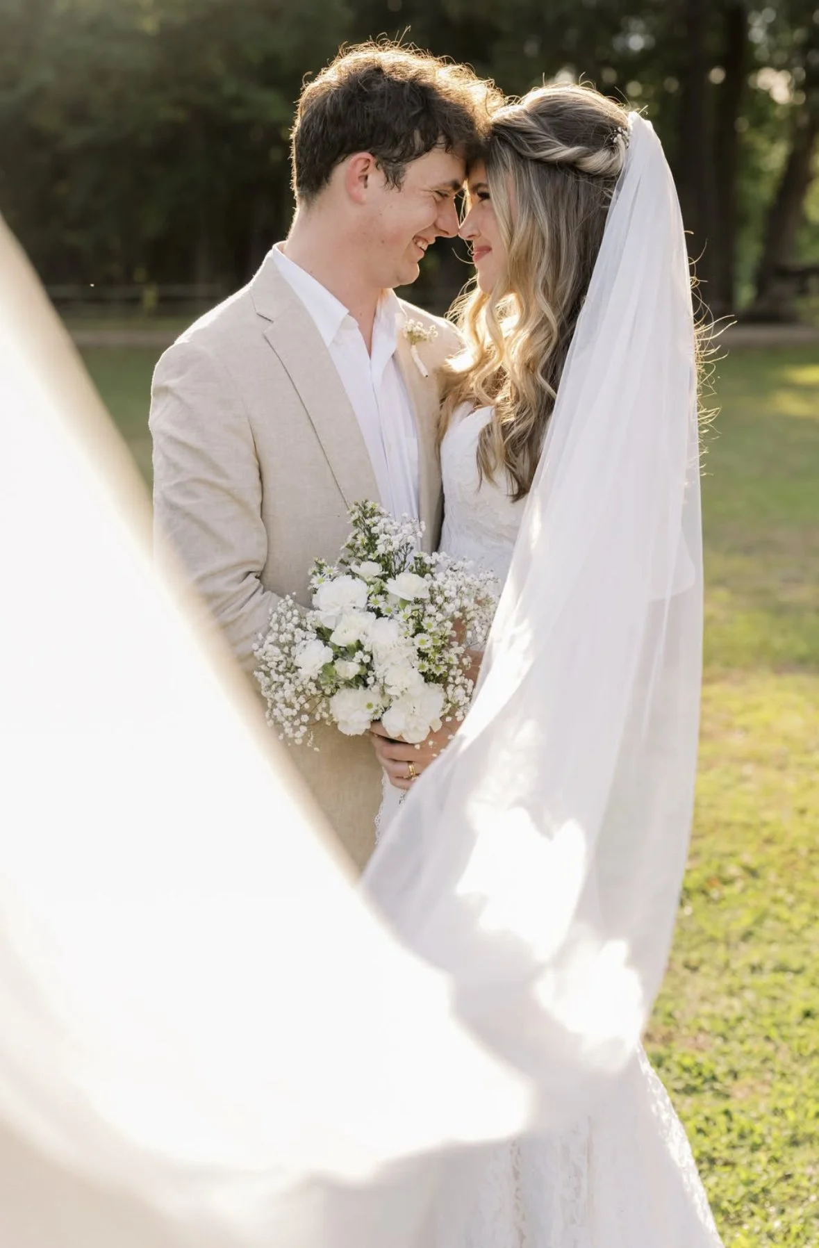 A bride and groom with their foreheads touching, smiling, outdoors during sunset, woman in white wedding dress with veil, man in beige suit, holding a bouquet of white flowers.