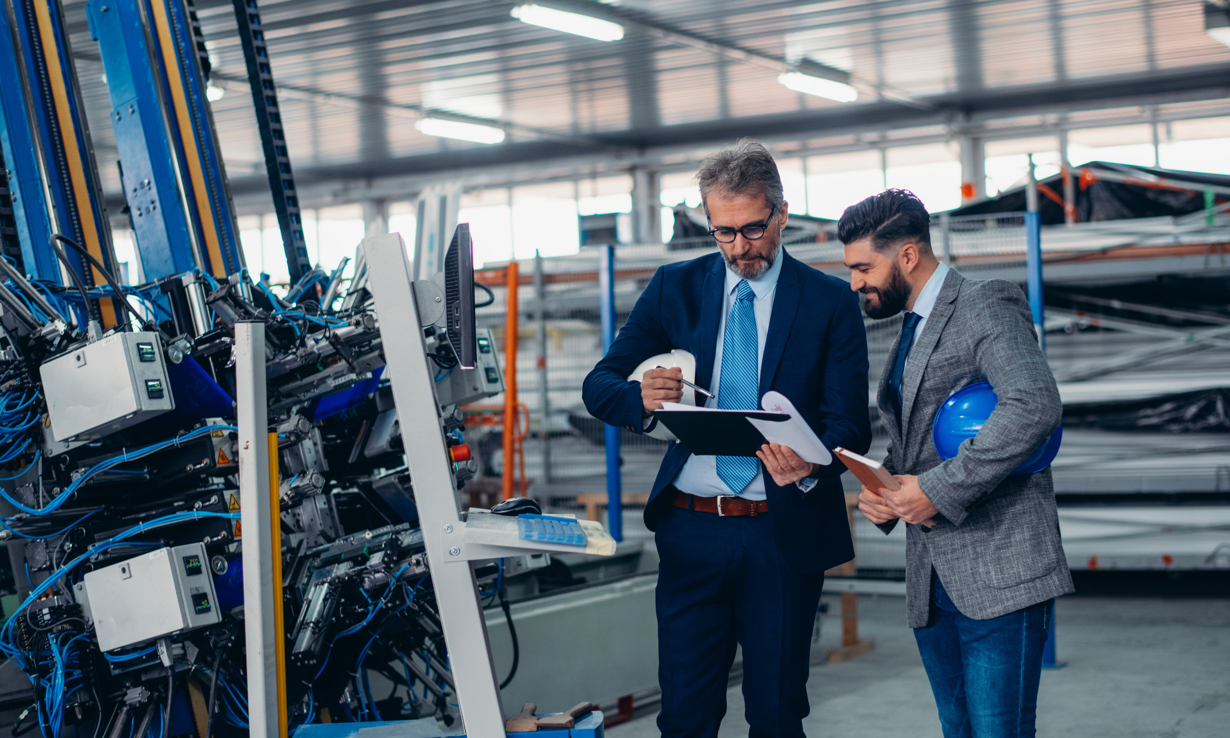 Two engineers in business suits and holding helmets, inspecting machinery in an industrial or manufacturing facility with shelves and equipment in the background.