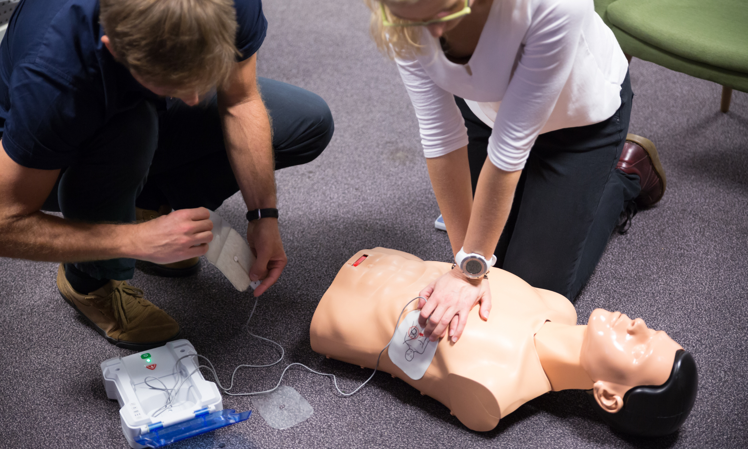 Man and Women doing a First Aid workshop - There is an 'Annie' Resuscitation doll on the floor. They are learning how to use a defibrillator machine