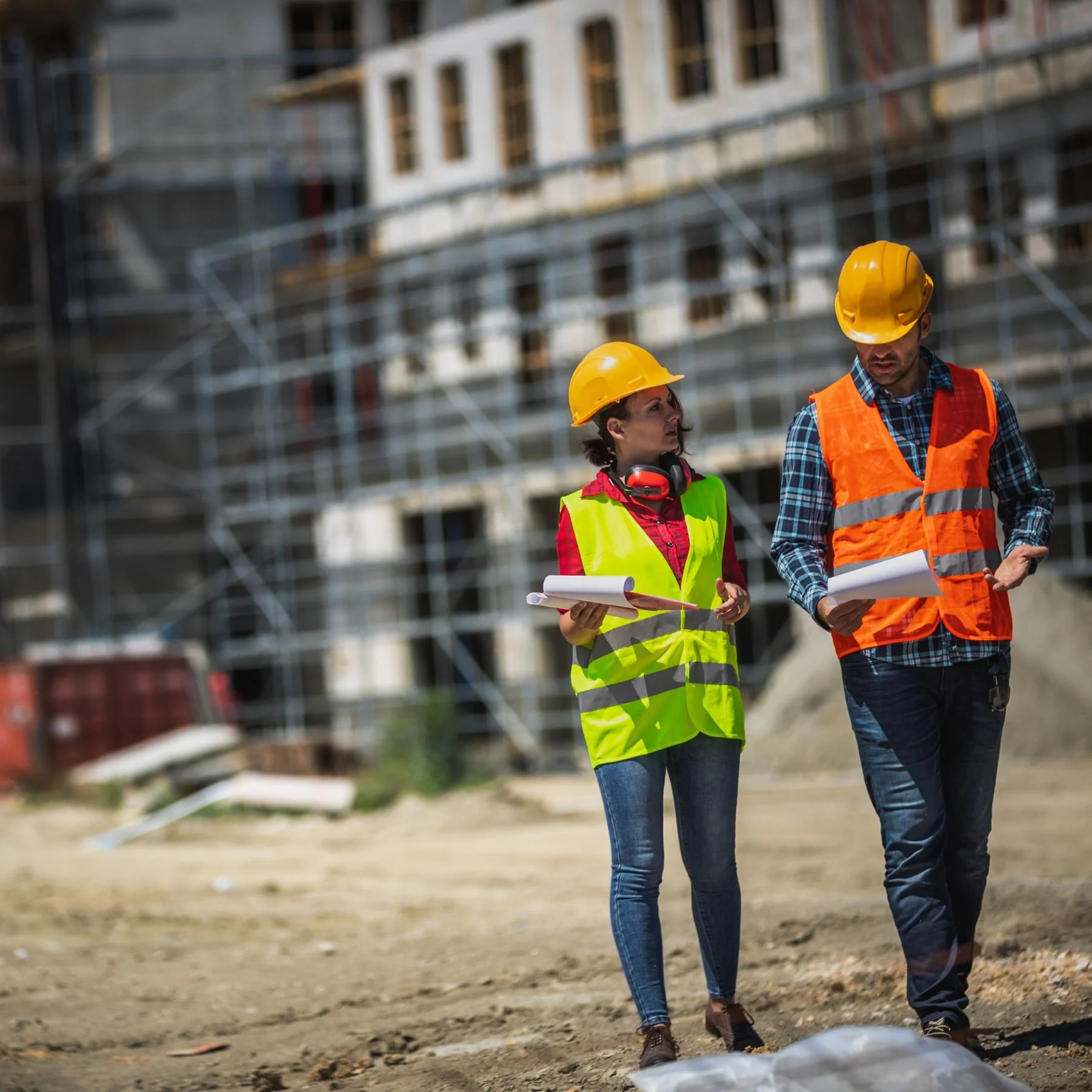 Two construction workers, a woman and a man, wearing yellow hard hats and safety vests, inspecting documents at a construction site with a building under construction in the background.