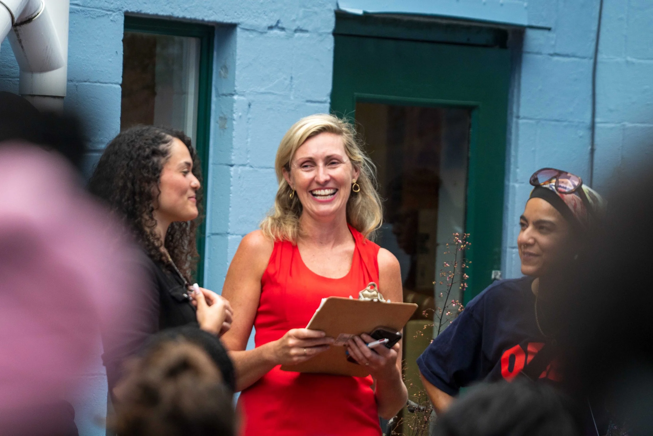 A woman in a red dress smiling and holding a clipboard, surrounded by a group of people in conversation, against a background of a blue brick wall and a green door.