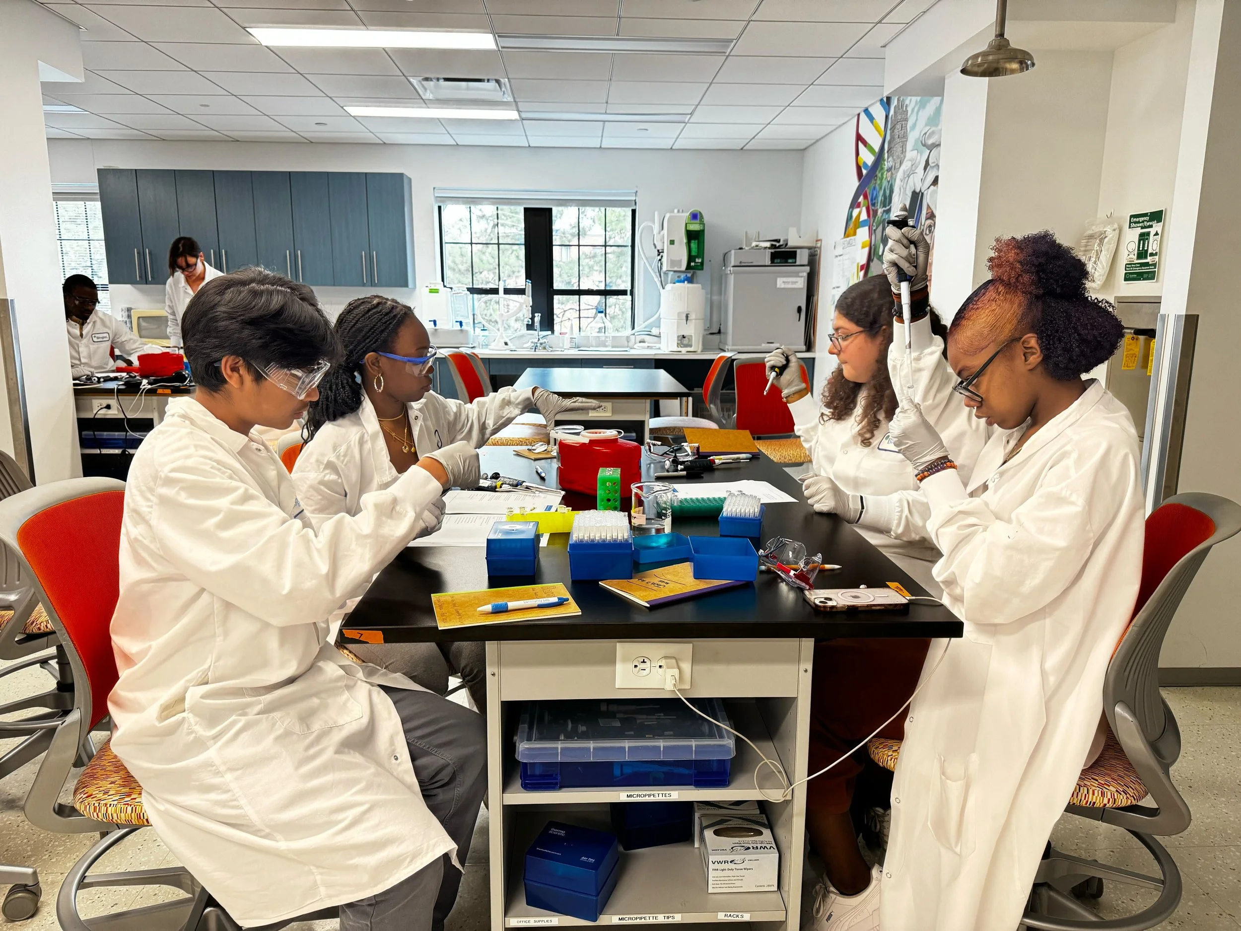 A group of five diverse students in white lab coats working together in a science classroom or laboratory, handling test tubes and equipment at a black lab table.