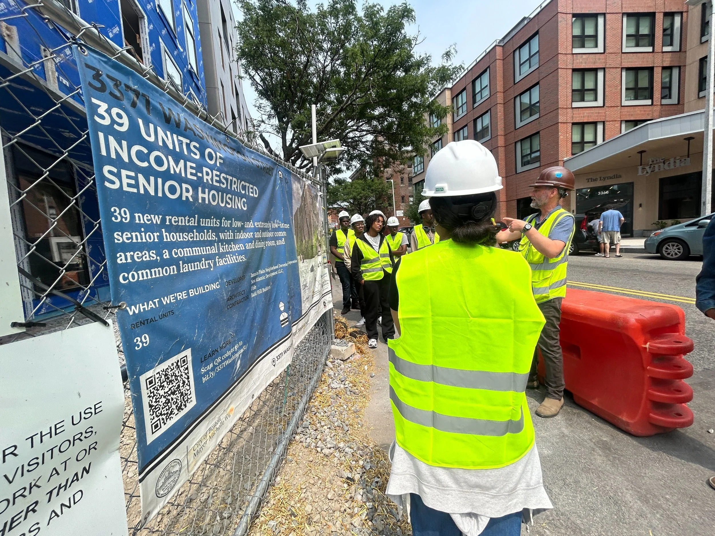 Construction workers wearing yellow safety vests and helmets gathered around a site sign on a city street, with buildings and parked cars in the background.