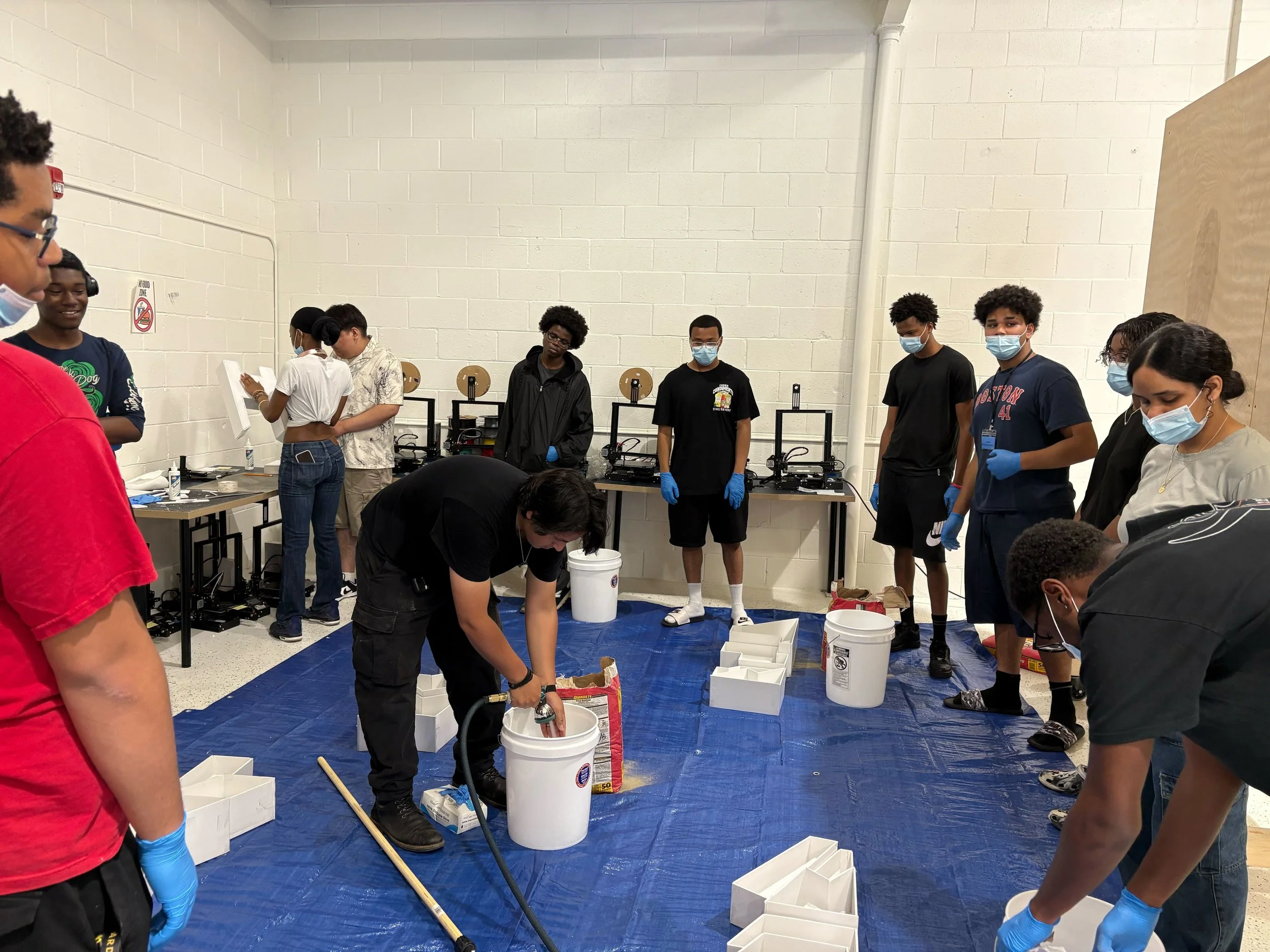 Group of diverse students and instructor in a classroom setting, wearing masks and gloves, engaging in a hands-on science activity involving buckets and materials on a blue tarp.