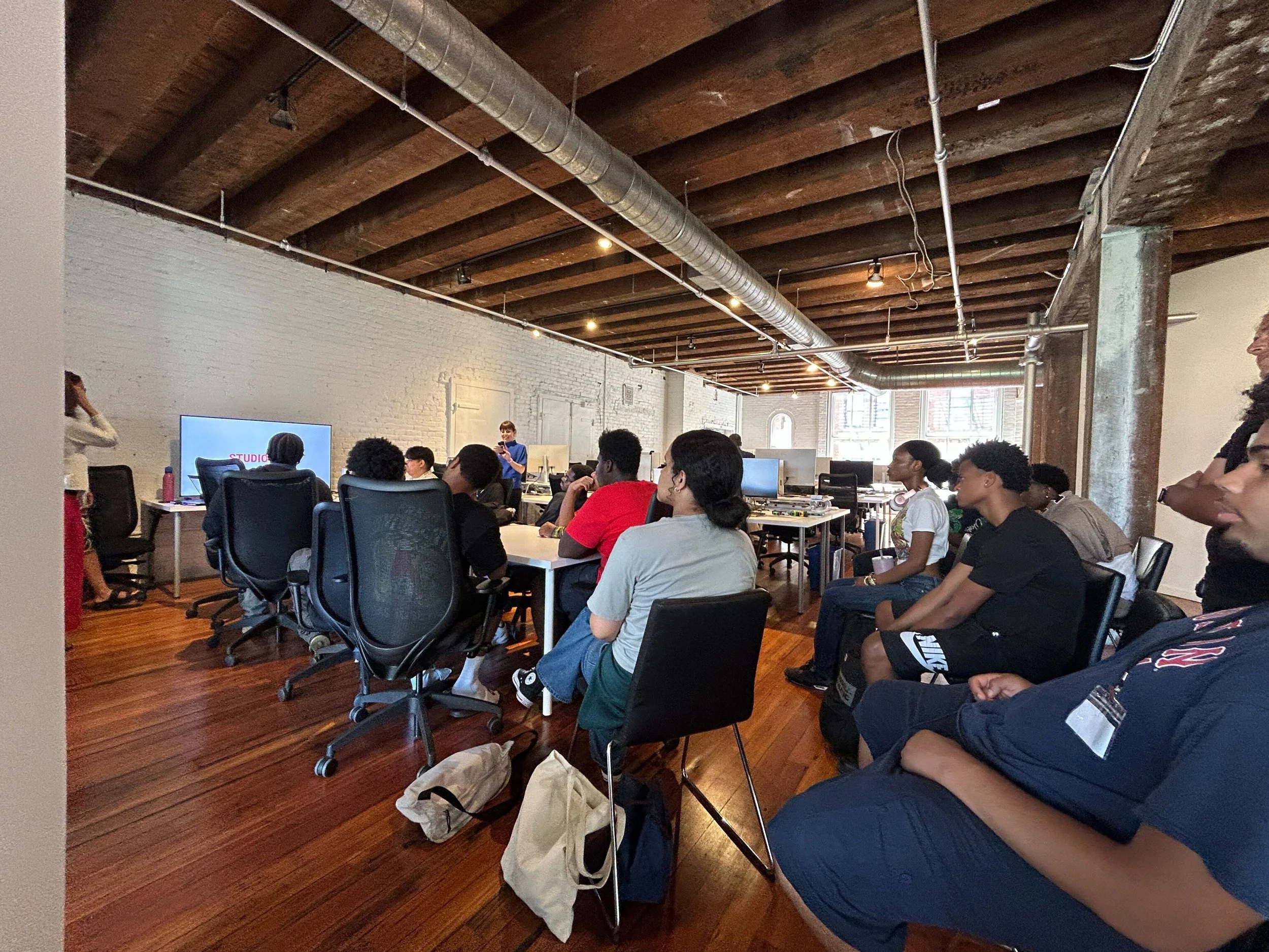 Several people seated in chairs and at tables in a spacious, industrial-style office or classroom with exposed brick walls, wooden beams, and ductwork, watching a person giving a presentation.