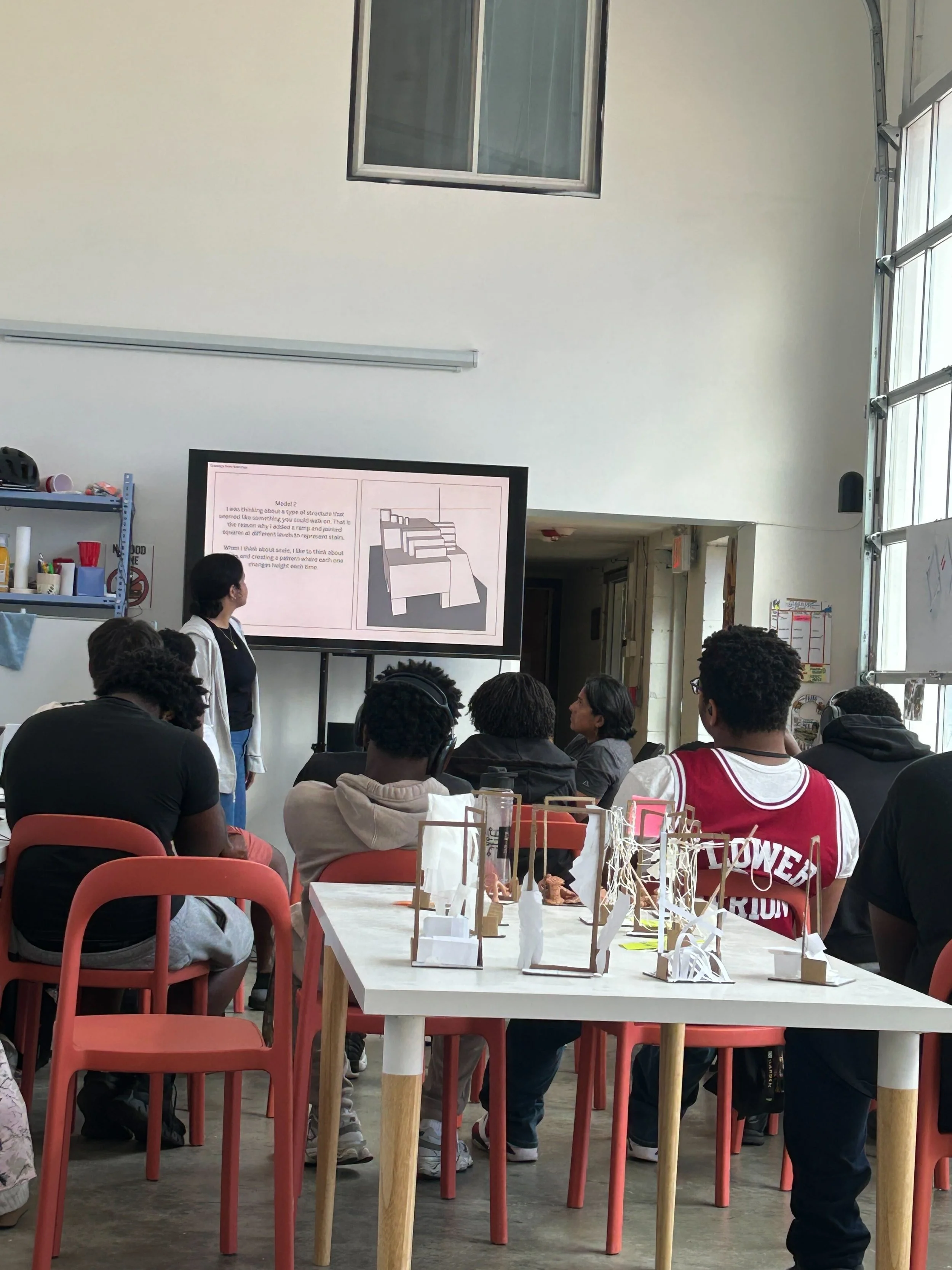 Students in a classroom watching a presentation on a monitor, with models of architectural structures on a table in the foreground.