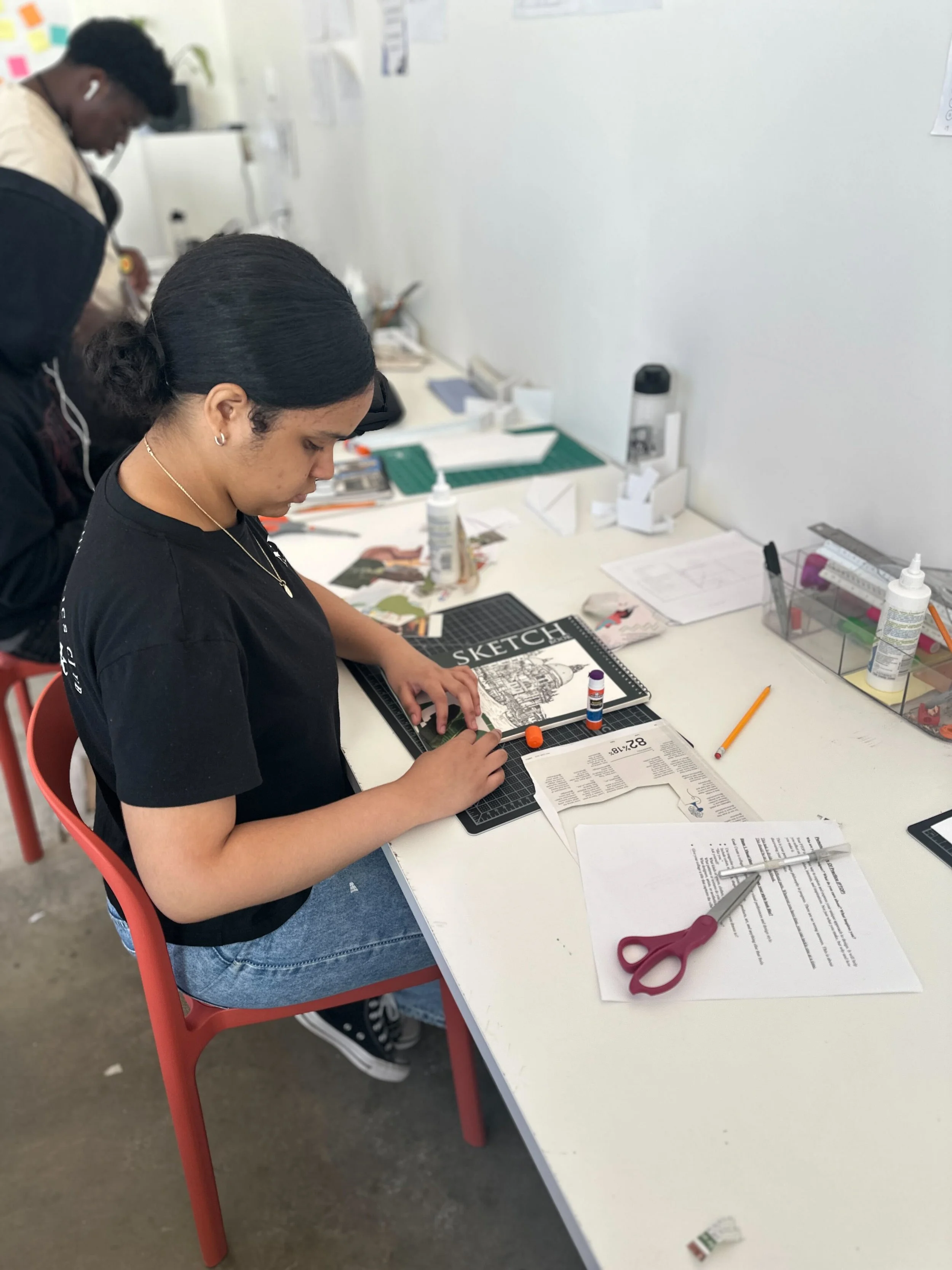 A young woman working at a desk in a classroom or office, using a paper craft project with glue stick and scissors, with other supplies and papers visible on the desk.