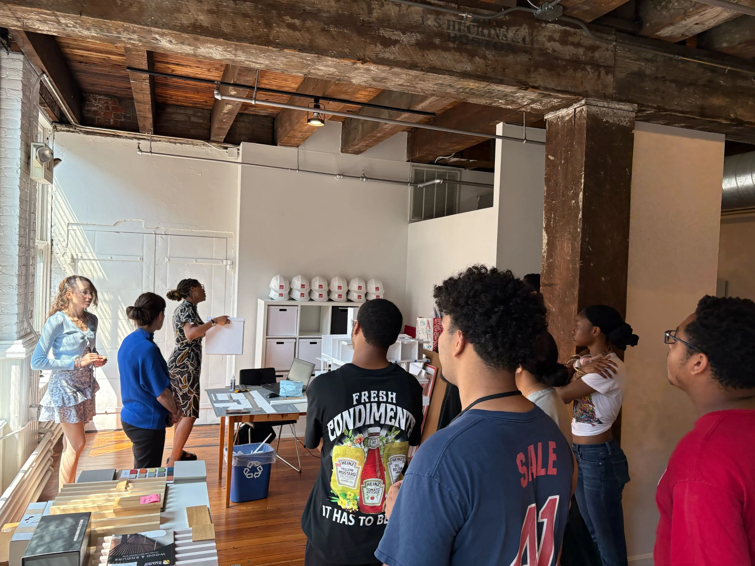 Group of people attending a presentation in an industrial-style room with exposed wooden beams, white walls, and natural light from large windows.