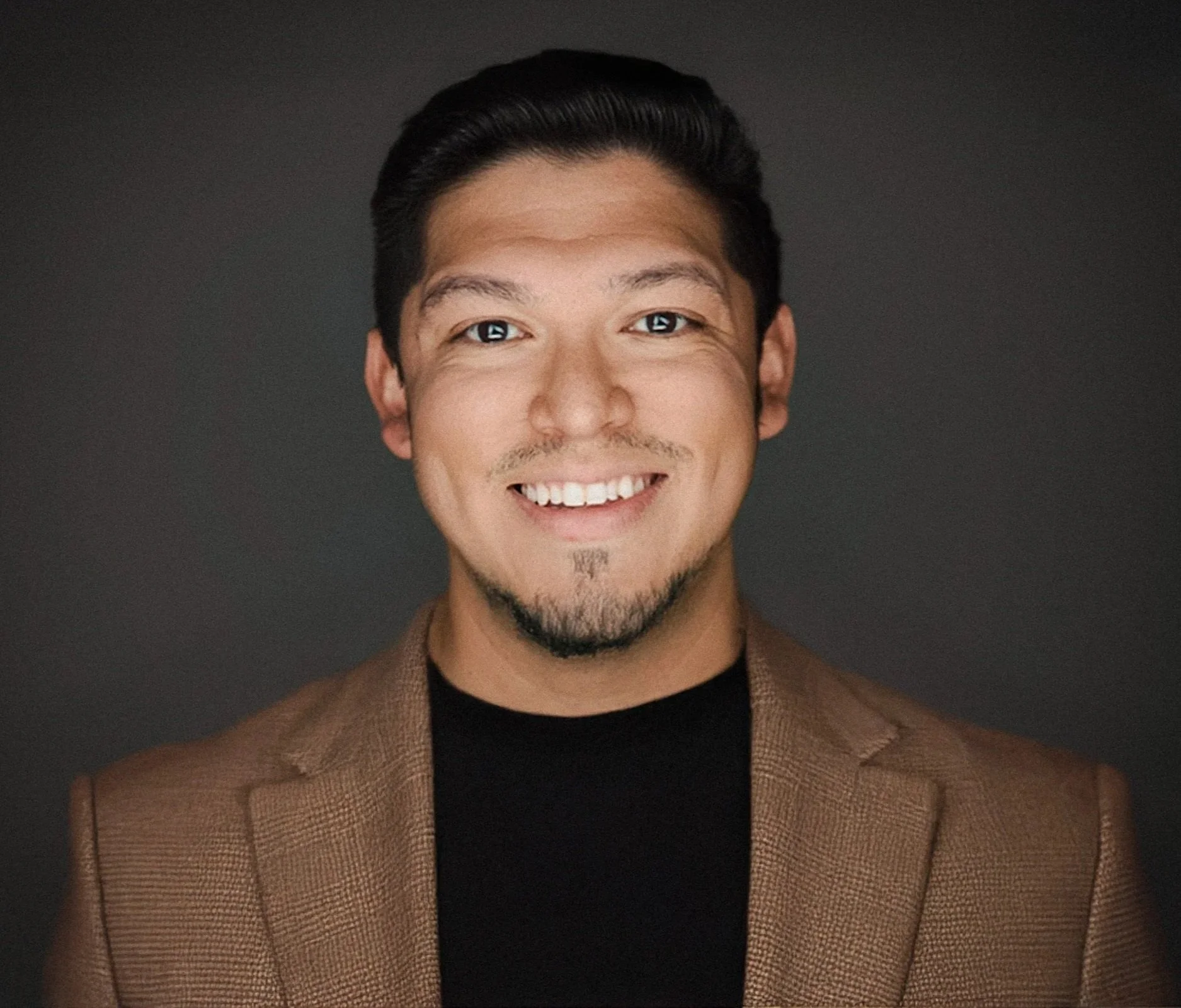 Portrait of a young man with dark hair, smiling, wearing a brown blazer and black shirt, against a dark background.