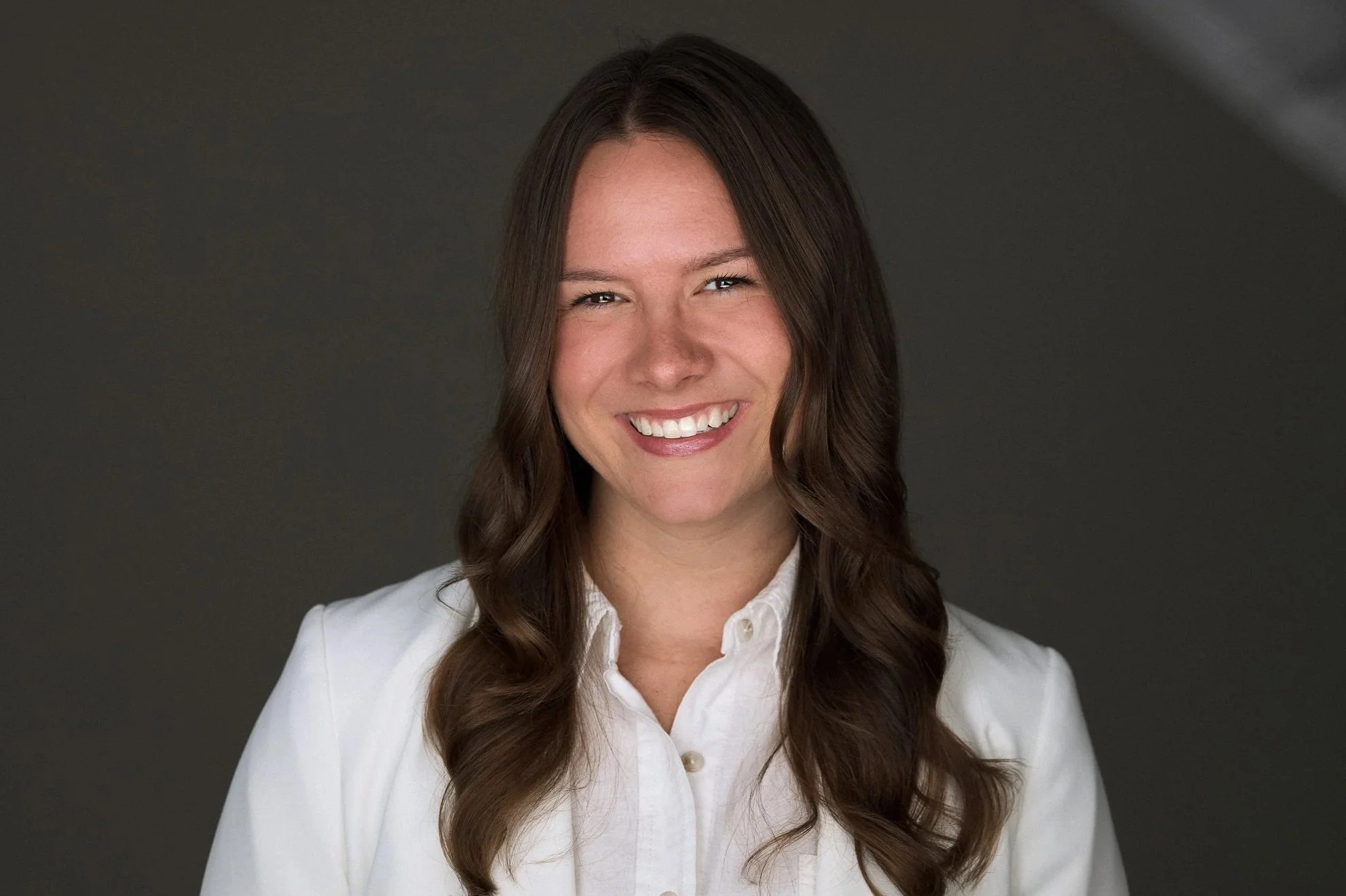 A woman with long brown hair smiling, wearing a white button-up shirt, against a dark background.