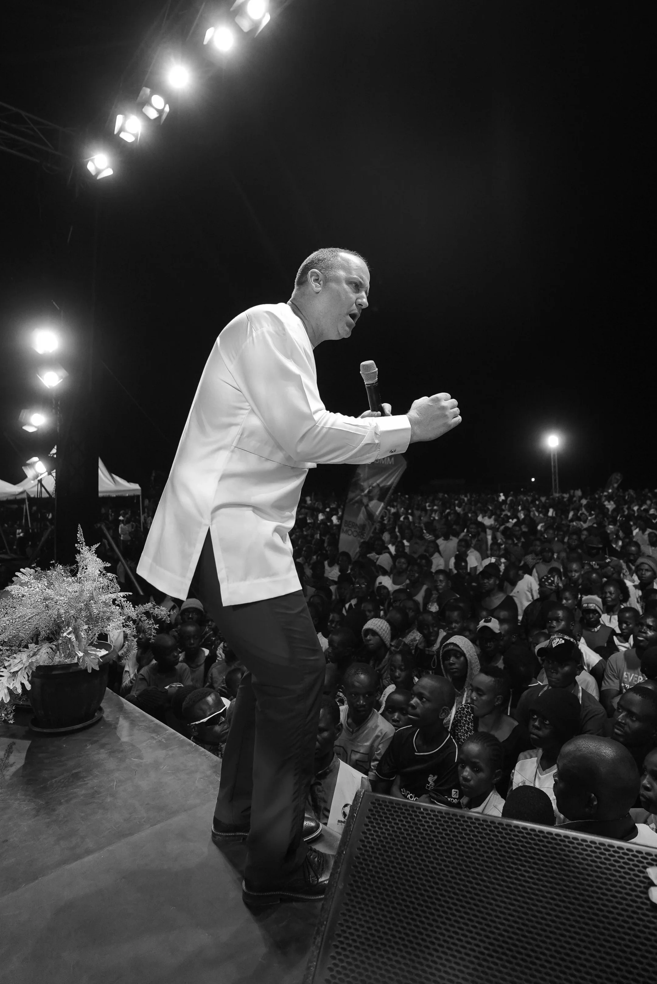 A man in a white shirt and dark pants speaking or shouting into a microphone on stage during a nighttime outdoor event with a large crowd in the audience.