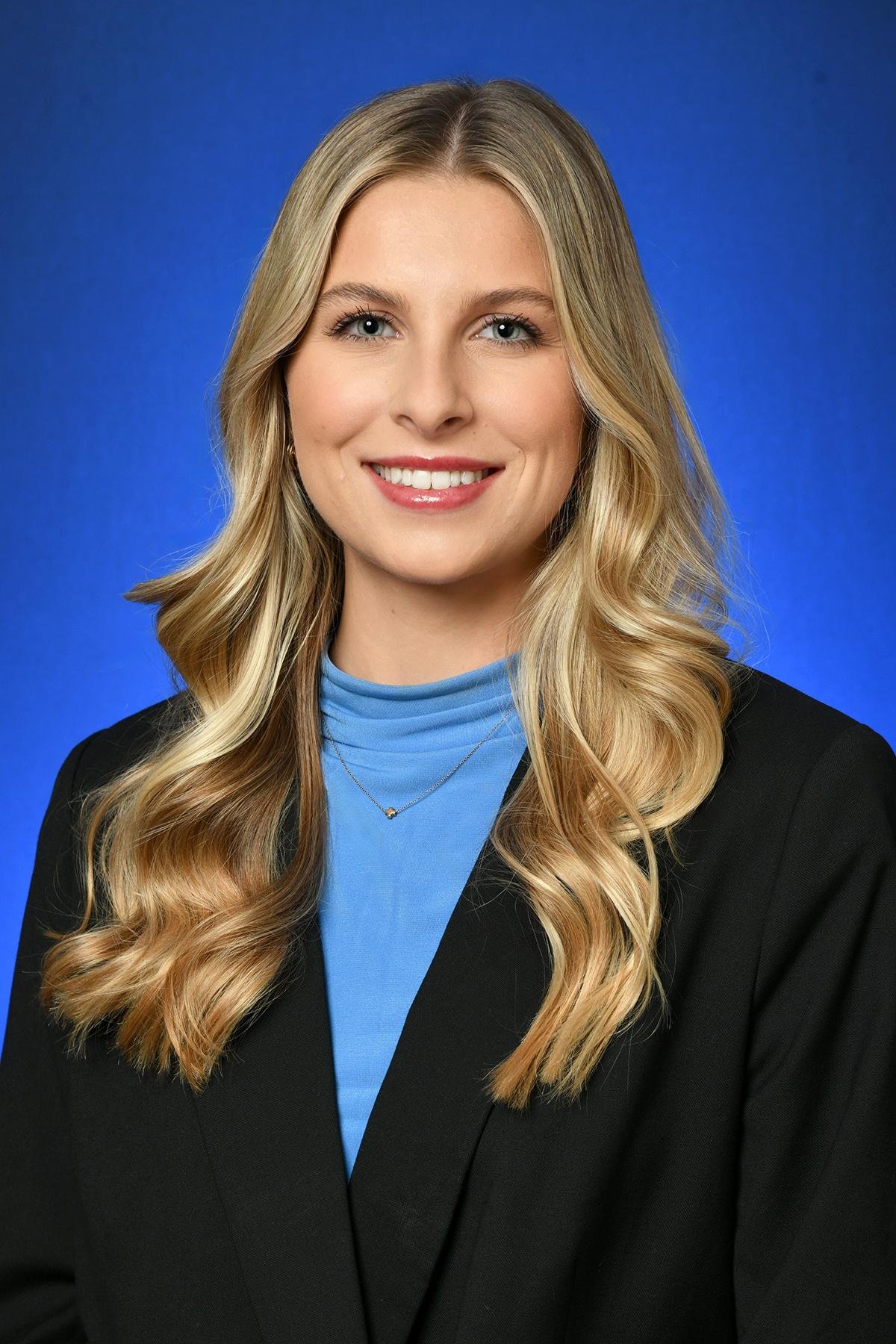 A woman with long blonde wavy hair wearing a blue shirt, black blazer, and a delicate necklace, smiling against a blue background.