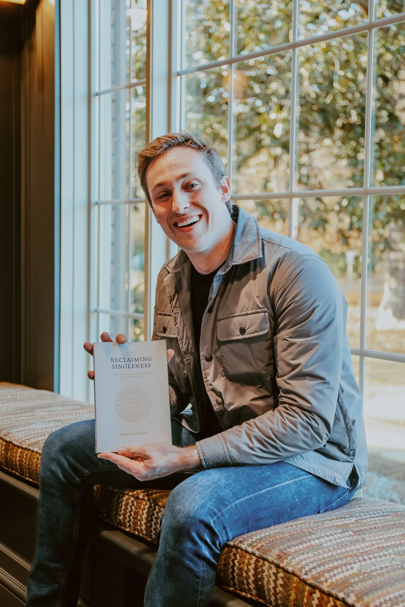 A young man with short brown hair, wearing a gray jacket and jeans, is sitting on a window seat with a colorful patterned cushion. He is smiling and holding a book titled "Reclaiming Singleness" by Sam McManus. The background shows a large window with trees and autumn leaves outside.