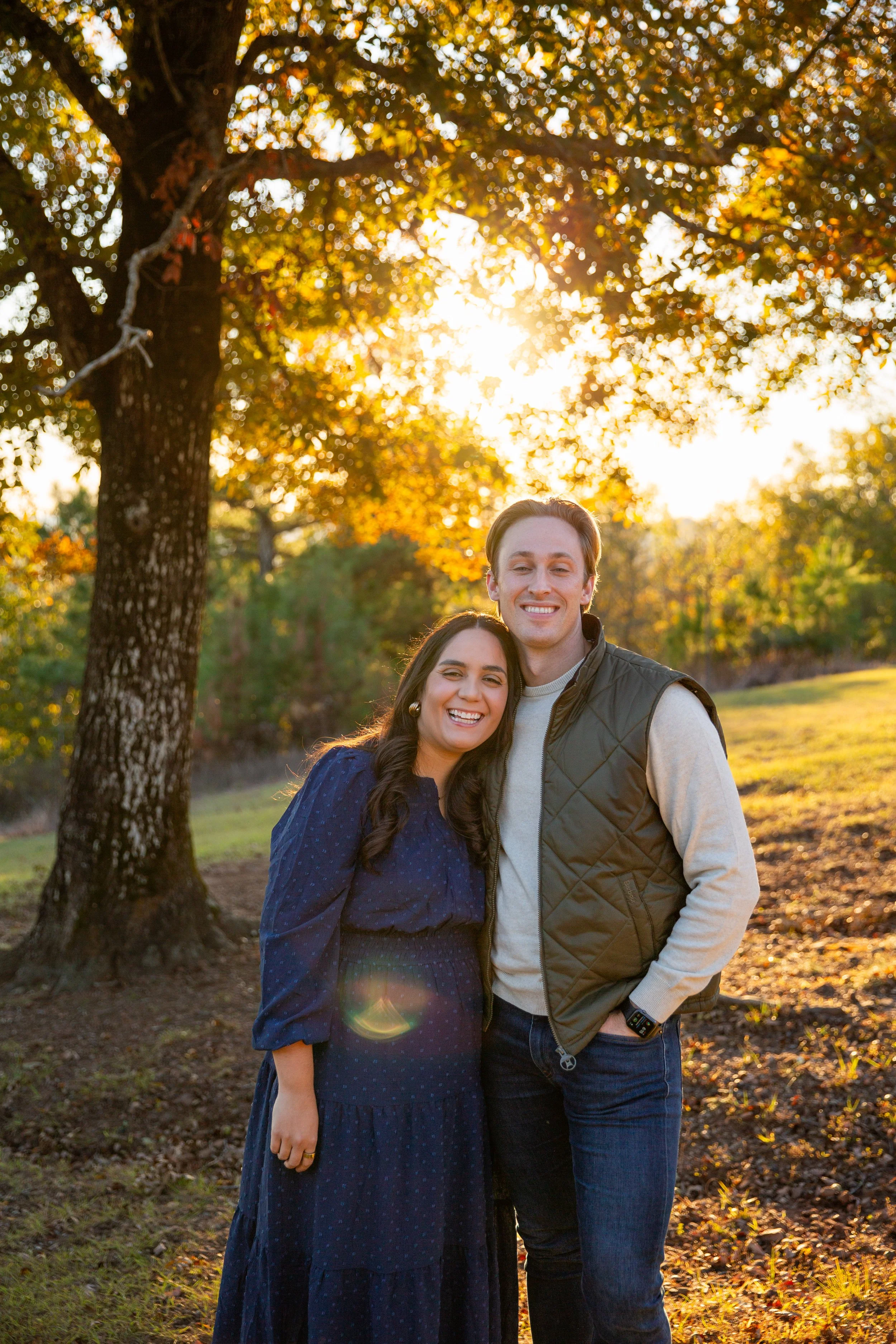 A smiling couple standing close together outdoors during sunset, in autumn, with a large tree and golden leaves and sunlight in the background.