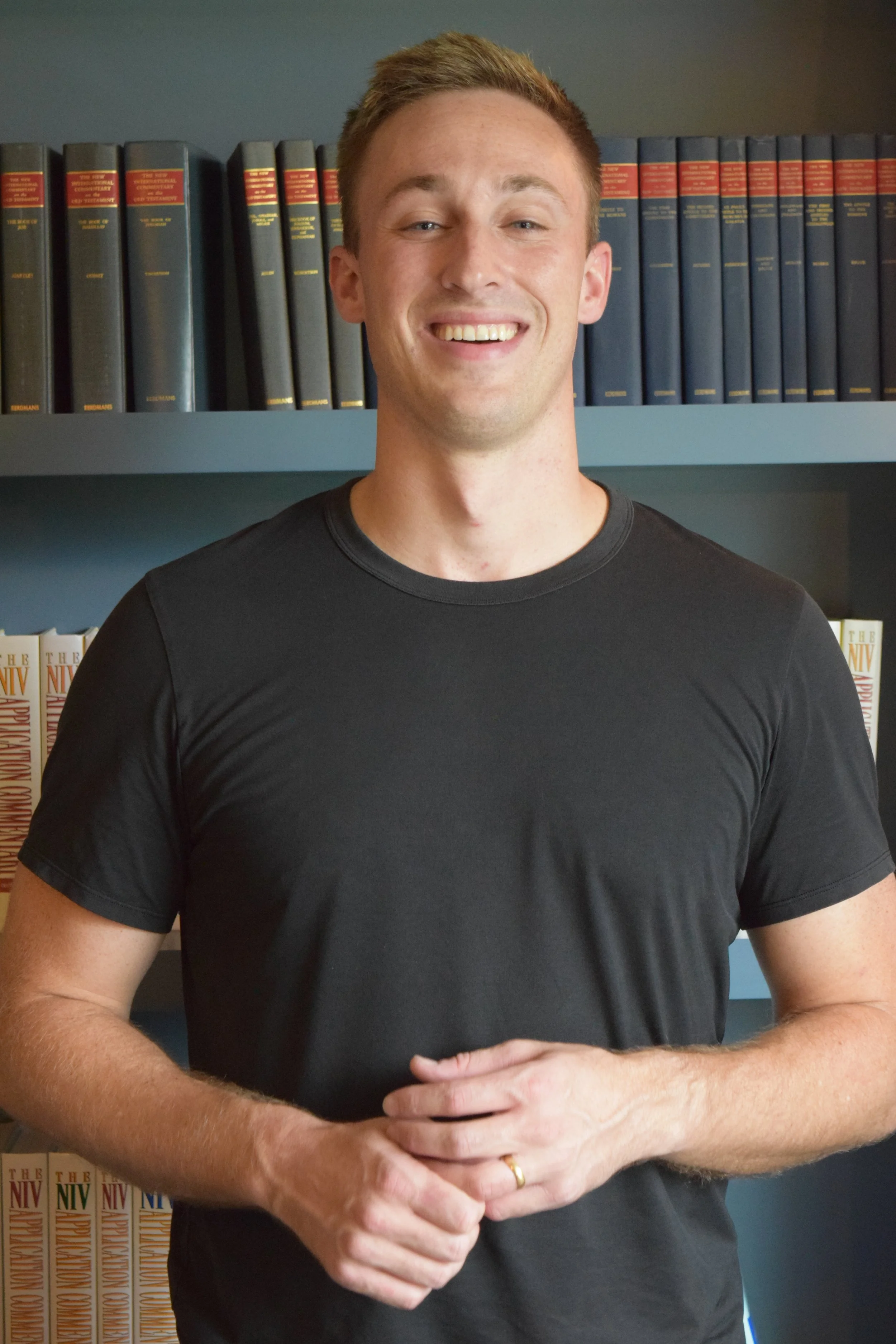 A smiling young man with short hair, wearing a black t-shirt, standing in front of a bookshelf with books, representing his coaching brand.