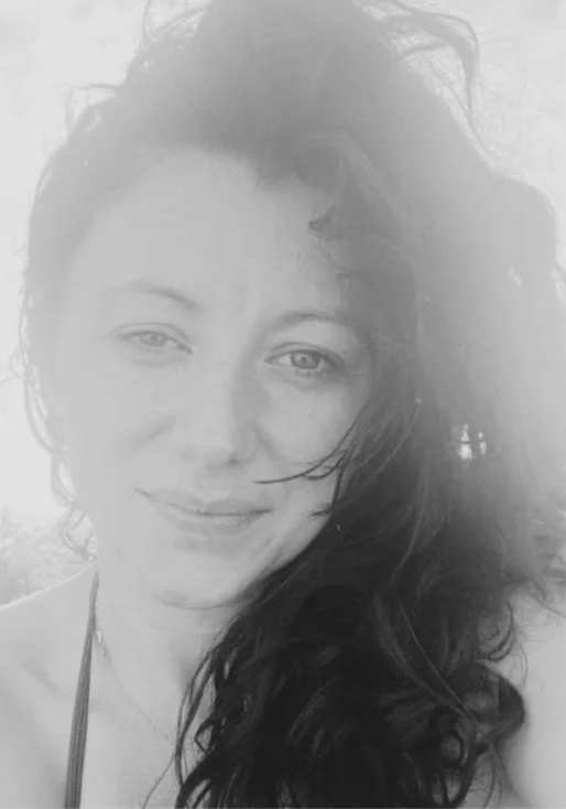 A woman with long, wavy hair smiling at the camera, outdoors with a cloudy sky.