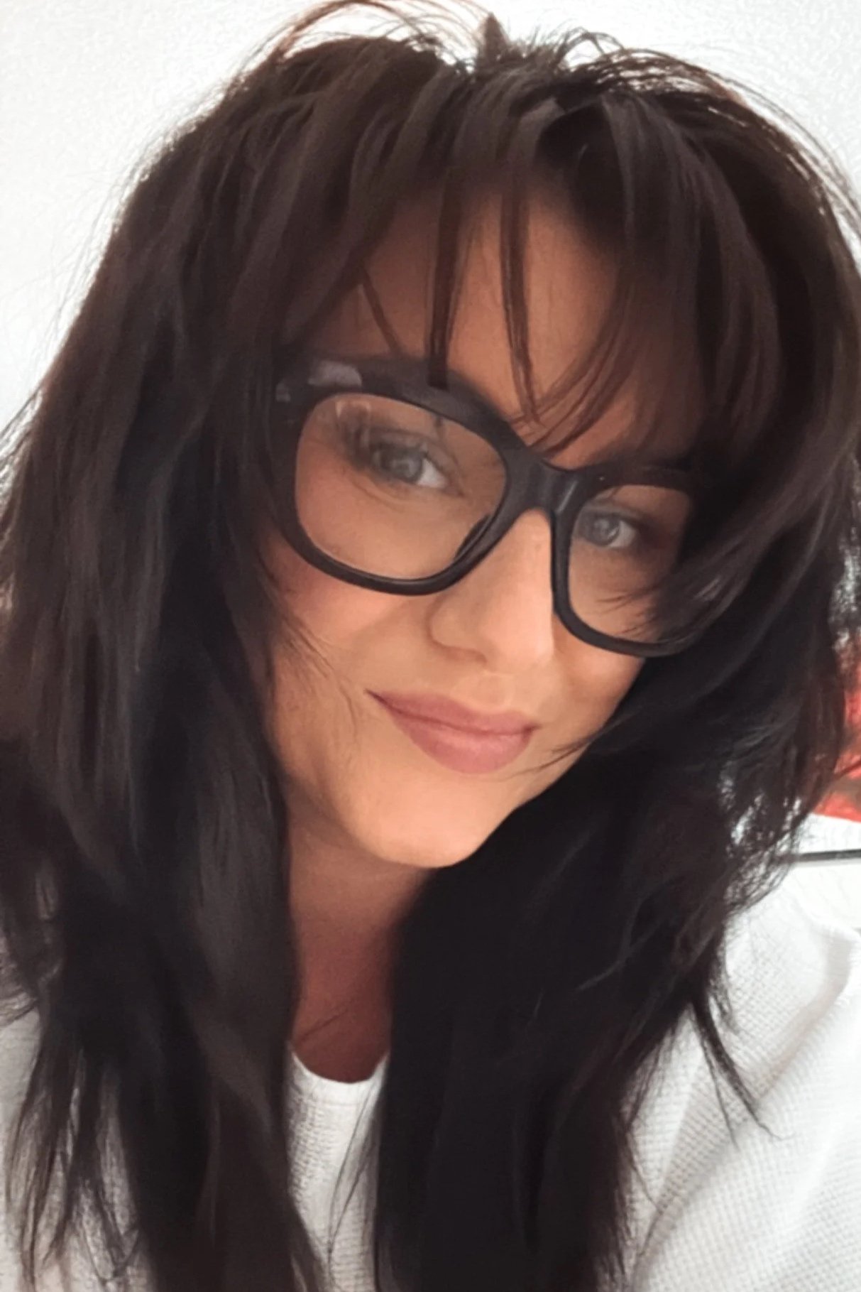 Close-up photo of a woman with dark hair, glasses, and a warm smile, wearing a white top.
