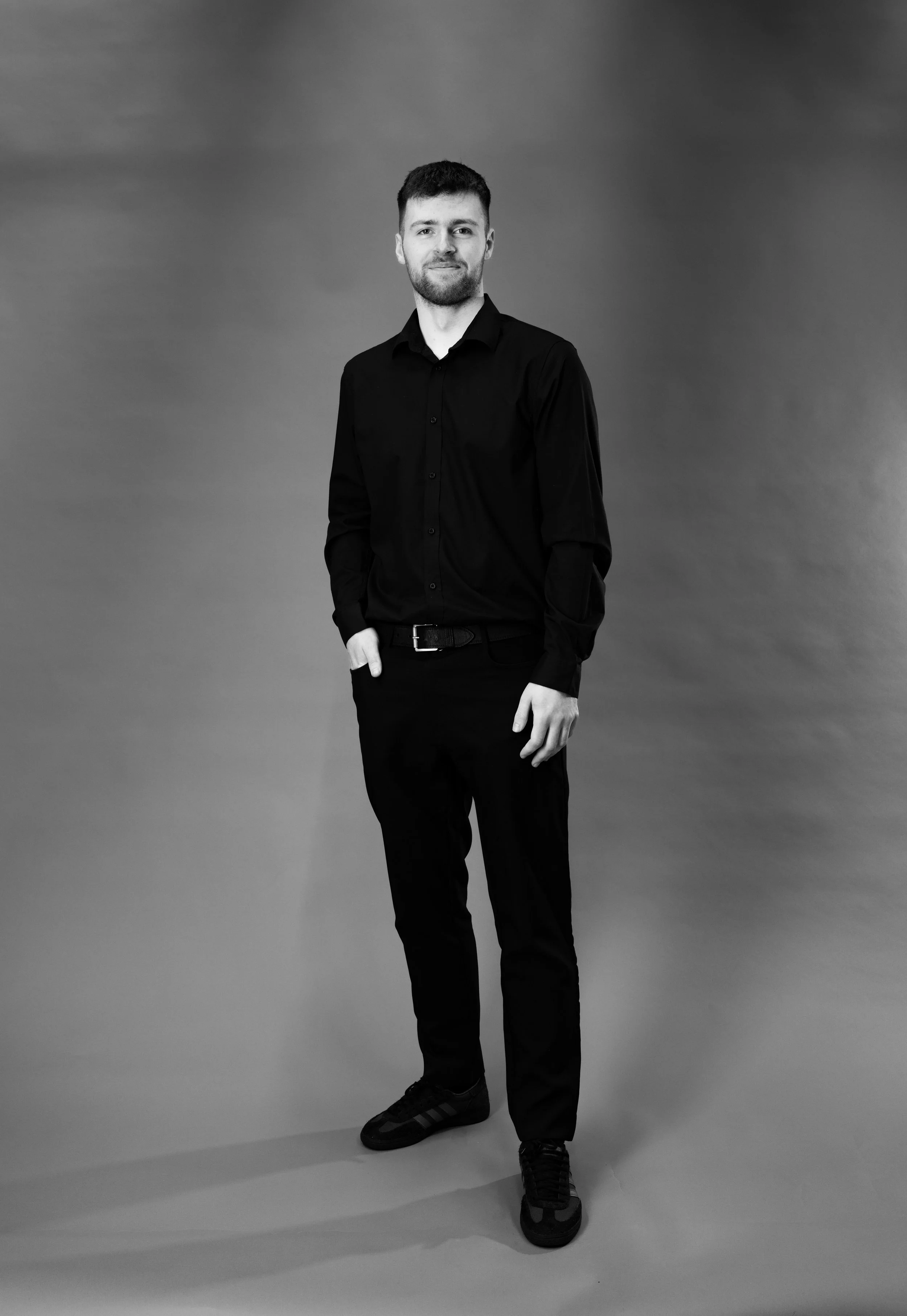 A young man with short hair and a beard wearing a black long-sleeve shirt, black pants, and sneakers stands against a plain background.