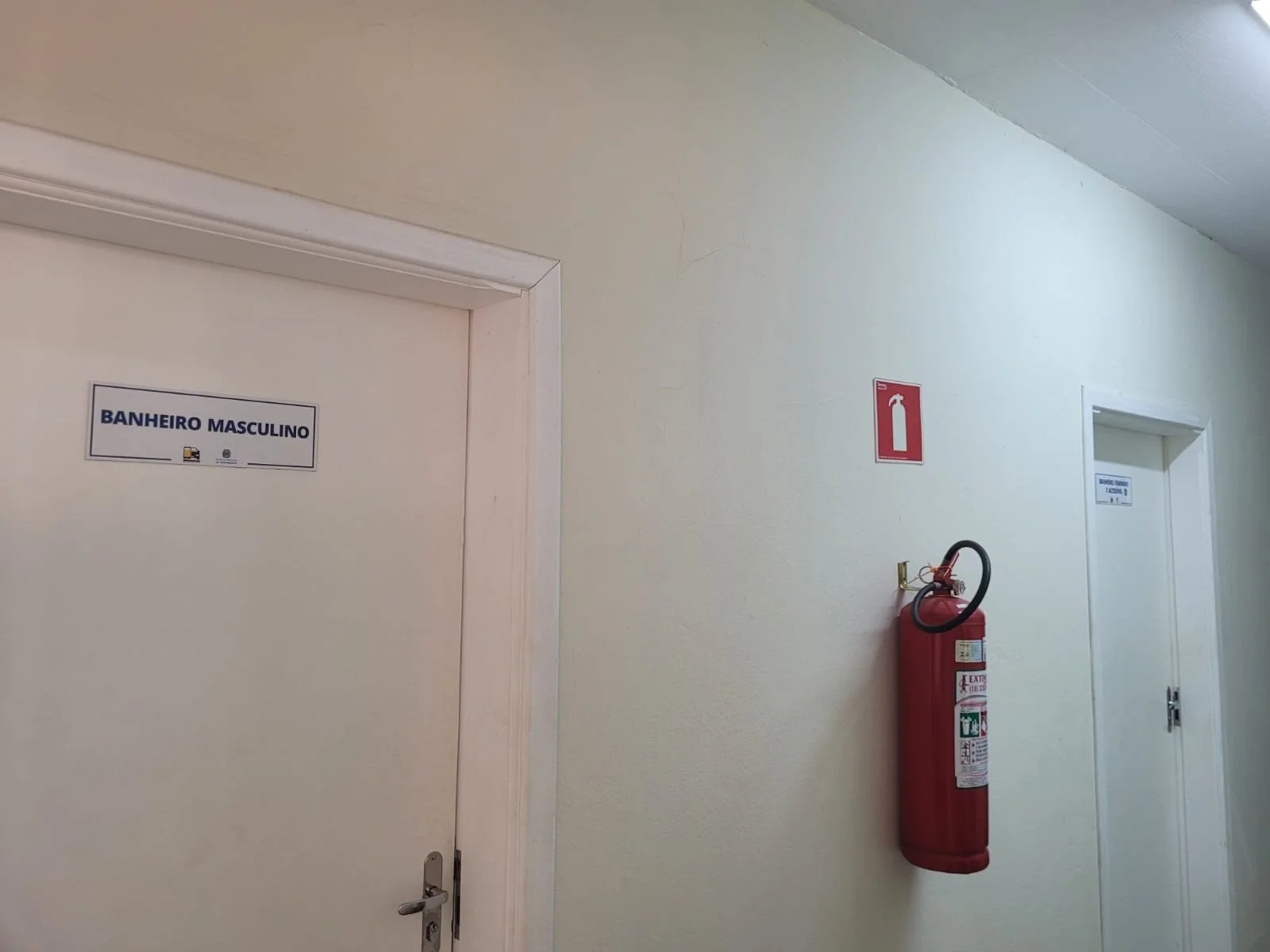Corredor com portas de banheiro masculino e extintor de incêndio vermelho na parede.