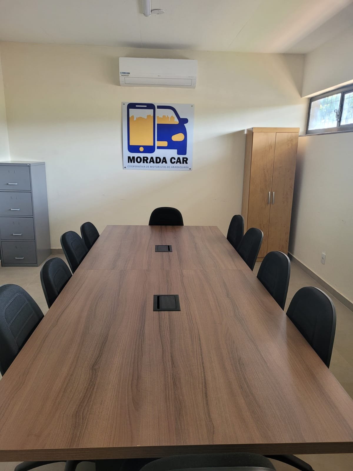 Sala de reuniões com mesa grande de madeira, cadeiras pretas, ar-condicionado e placa da Morada Car na parede.