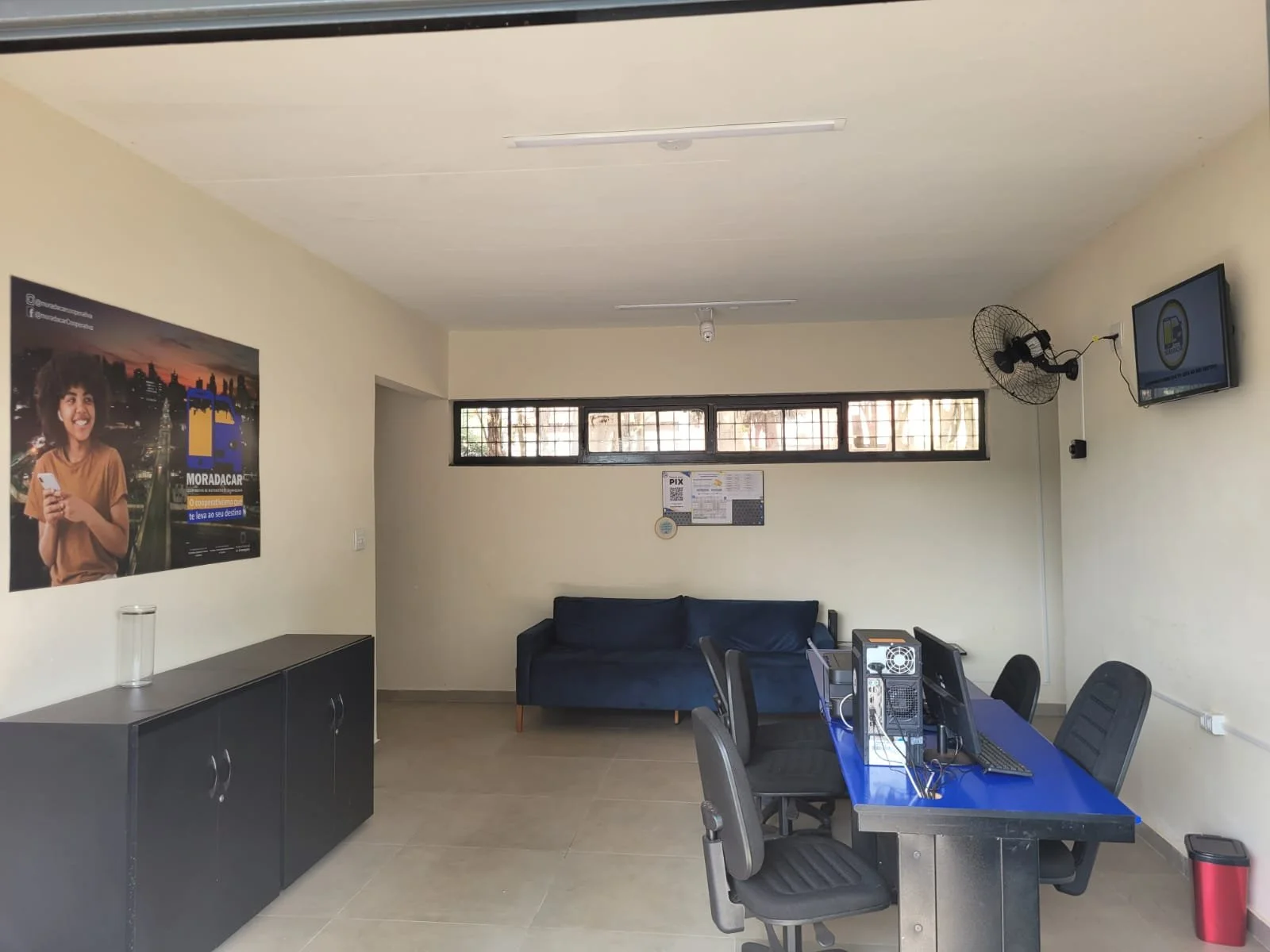 Sala de atendimento com sofá azul, mesa de computadores, cadeiras, armário, ventilador de parede e cartazes informativos.